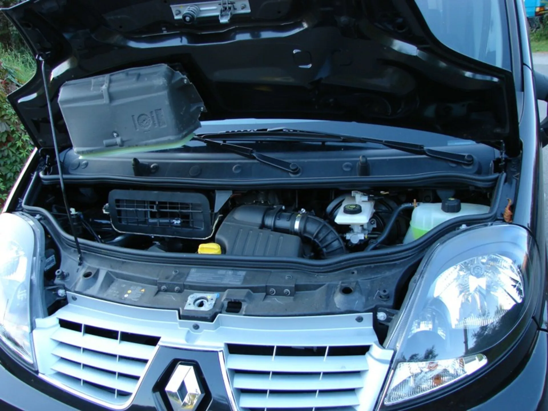 Vue avant trois quarts d'un Renault Trafic Combi 2006 avec capot ouvert révélant le compartiment moteur et composants, y compris bloc moteur avec couvercle, admission d'air et réservoirs de liquides, flanqué de phares halogènes traditionnels