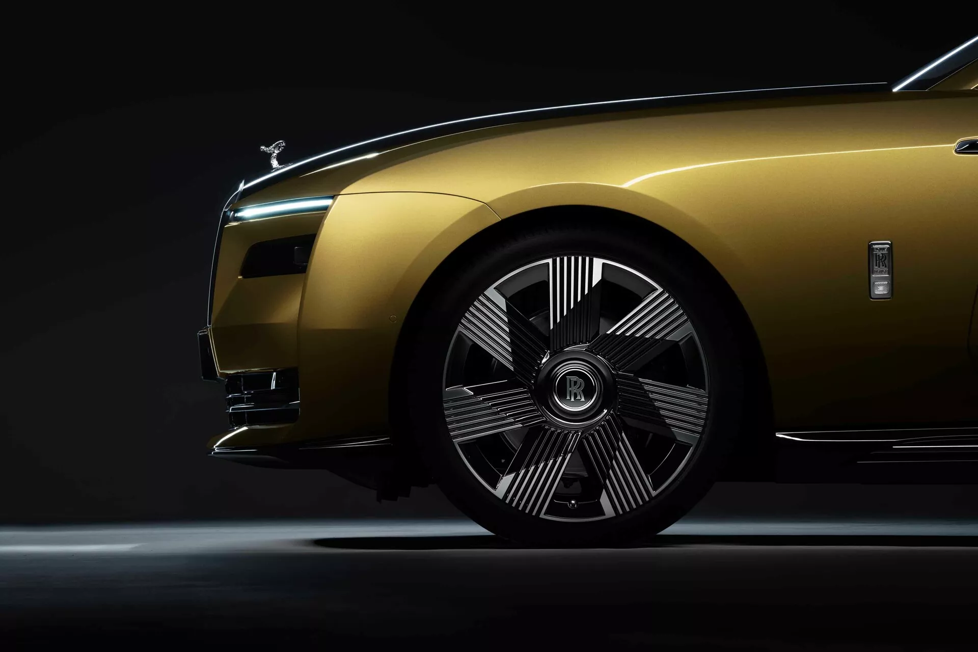 Vue de côté rapprochée et en contre-plongée d'une Rolls-Royce Spectre 2023 mettant en avant la roue avant, l'aile et une partie du capot sur fond sombre avec un éclairage dramatique pour souligner les caractéristiques de design et les courbes du véhicule de luxe.