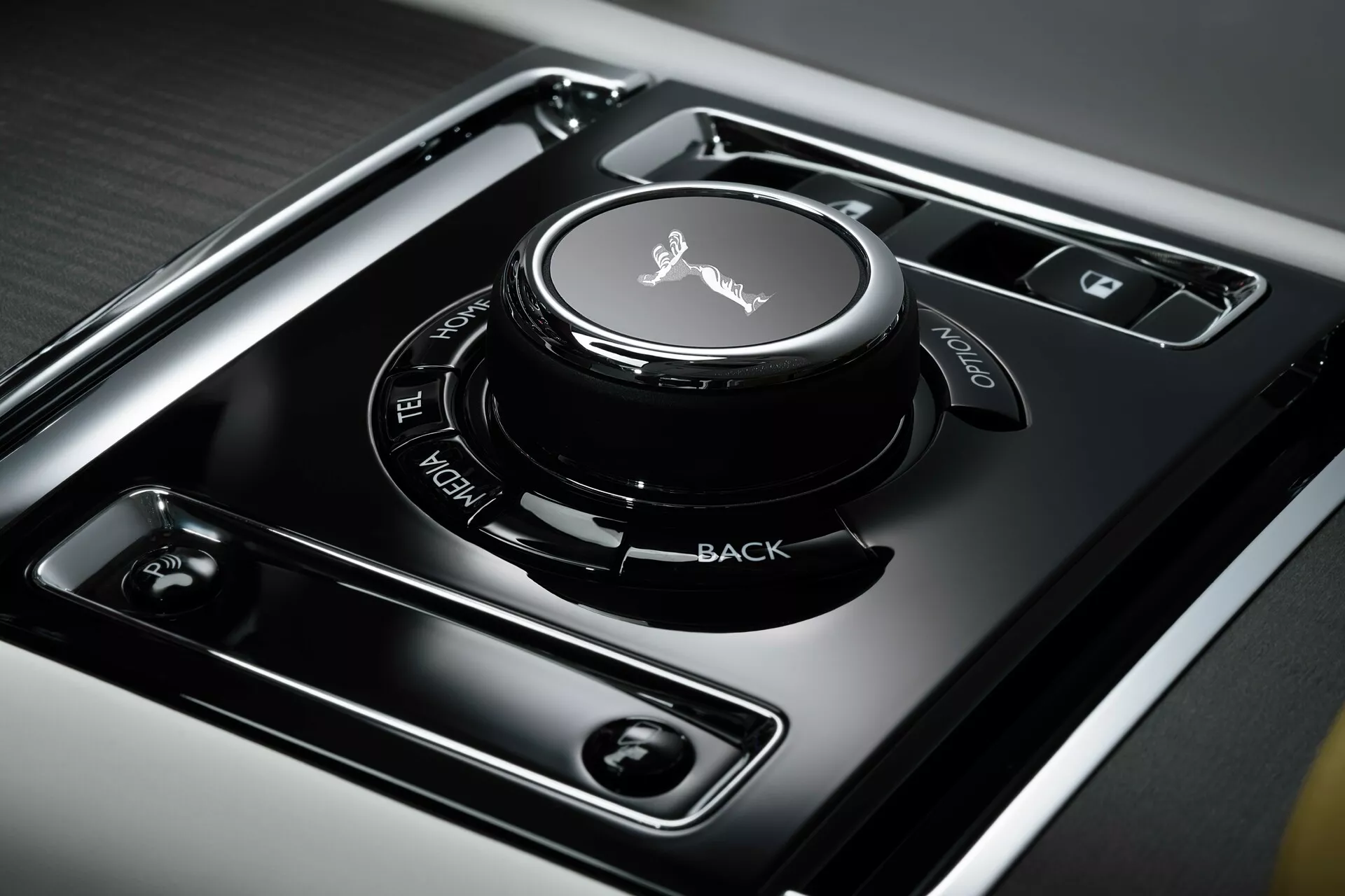 Vue rapprochée de la console centrale de la Rolls-Royce Spectre 2023 première génération, montrant le bouton de contrôle de l'infodivertissement avec une surface tactile, entouré de boutons aux fonctions clairement étiquetées sur un panneau noir brillant, situé dans un métal brossé pour un design intérieur luxueux