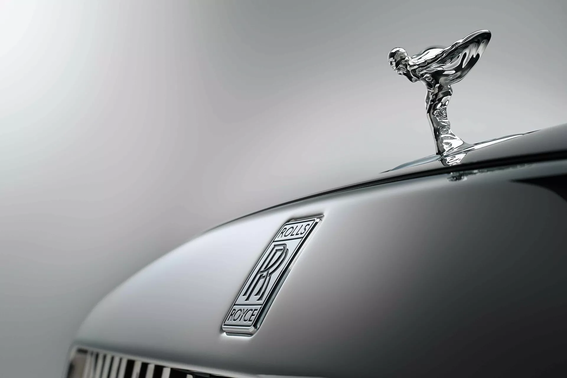 Vue rapprochée du capot avant de la Rolls-Royce Spectre 2023 avec une mise au point précise sur l'emblème Spirit of Ecstasy et la grille verticale luxueuse, définie contre un arrière-plan flou soulignant l'artisanat distingué de la voiture et sa finition gris foncé.