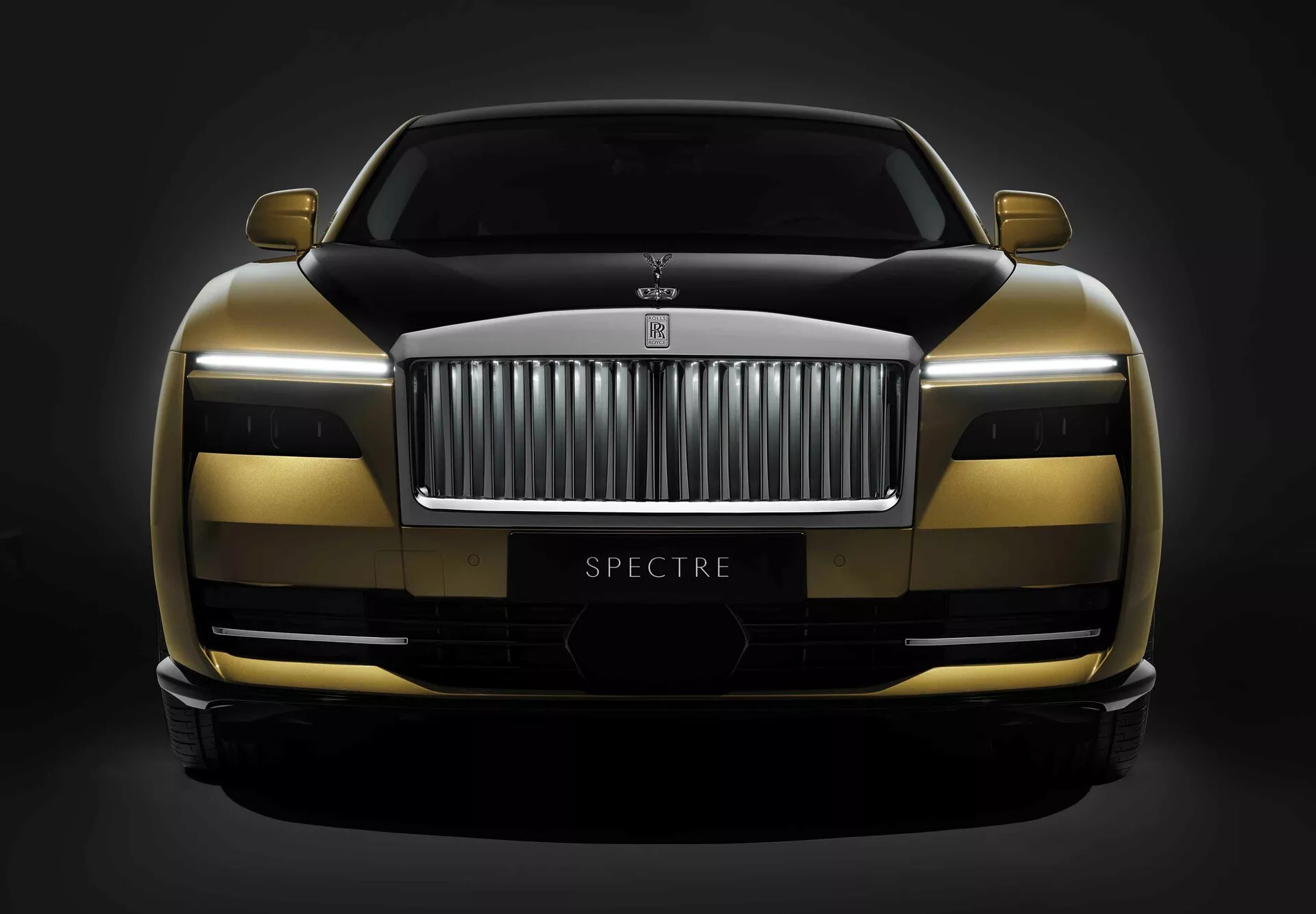 Rolls-Royce Spectre 1ère génération 2023 présentant un schéma de peinture bicolore, une technologie de phares avancée LED et un design profilé avec le nom du modèle 'SPECTRE' visible, capturée depuis un angle frontal bas sur un fond sombre.