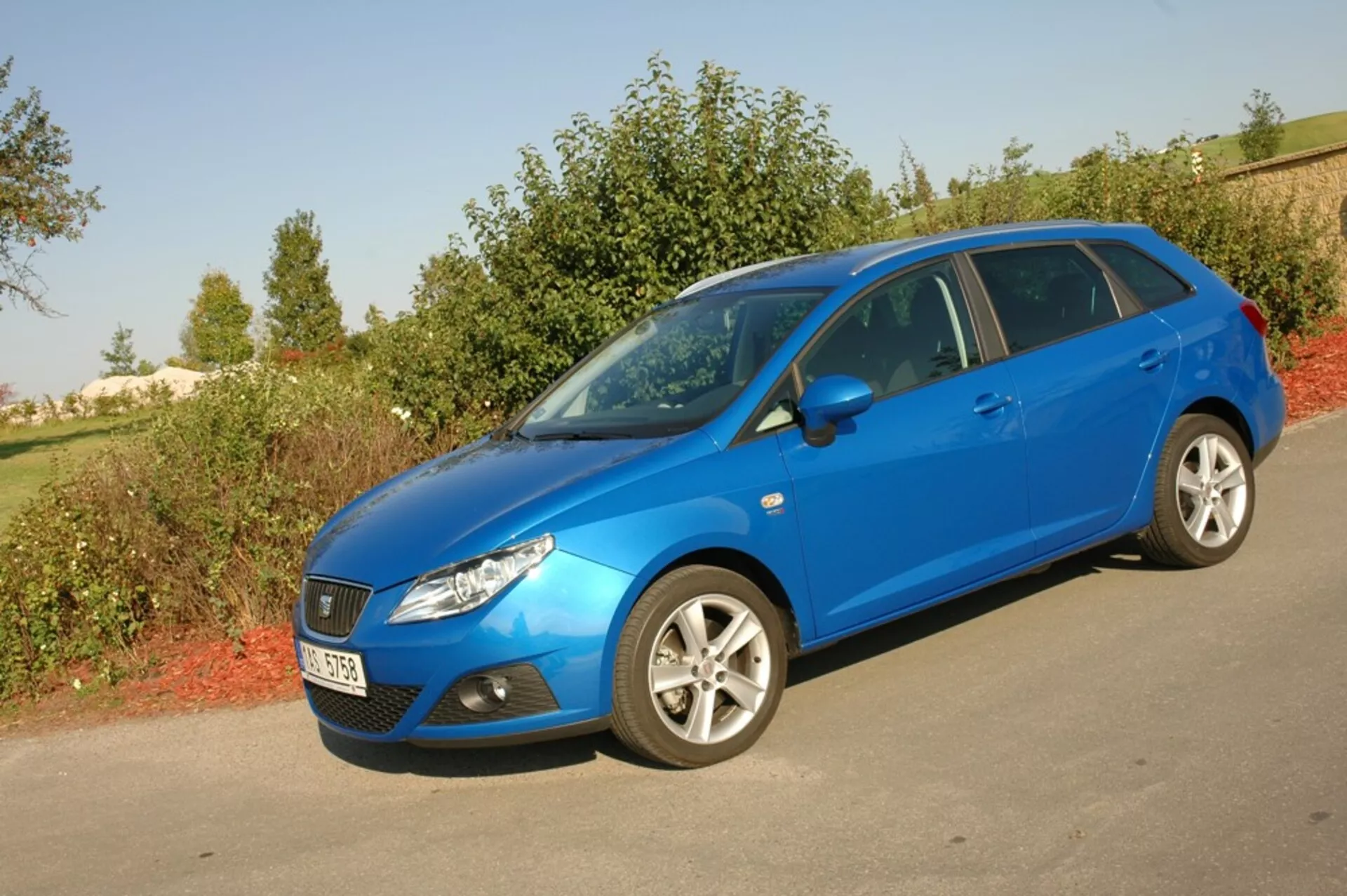 Seat Ibiza 2012 hatchback bleu vif garée sur le bitume face à la verdure, présentant sa grille avant aérodynamique, ses phares inclinés vers l'arrière et son profil latéral avec jantes en alliage multi-branches sous la lumière naturelle du jour