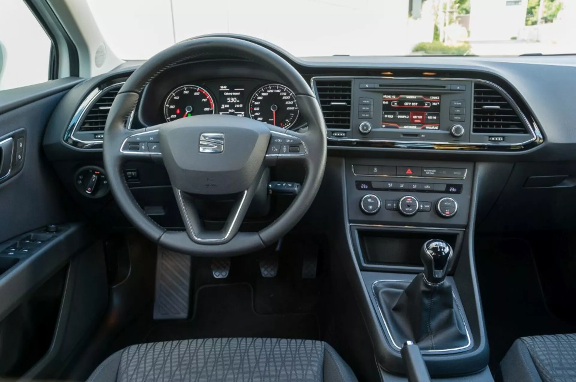 Vue intérieure d'une Seat Leon SC de 2016 depuis la perspective du siège passager avant, montrant le volant avec le logo du fabricant et les commandes montées, le tableau de bord numérique, le système d'infodivertissement, les commandes climatiques, le levier de vitesse manuel, et la porte du conducteur avec options électriques.