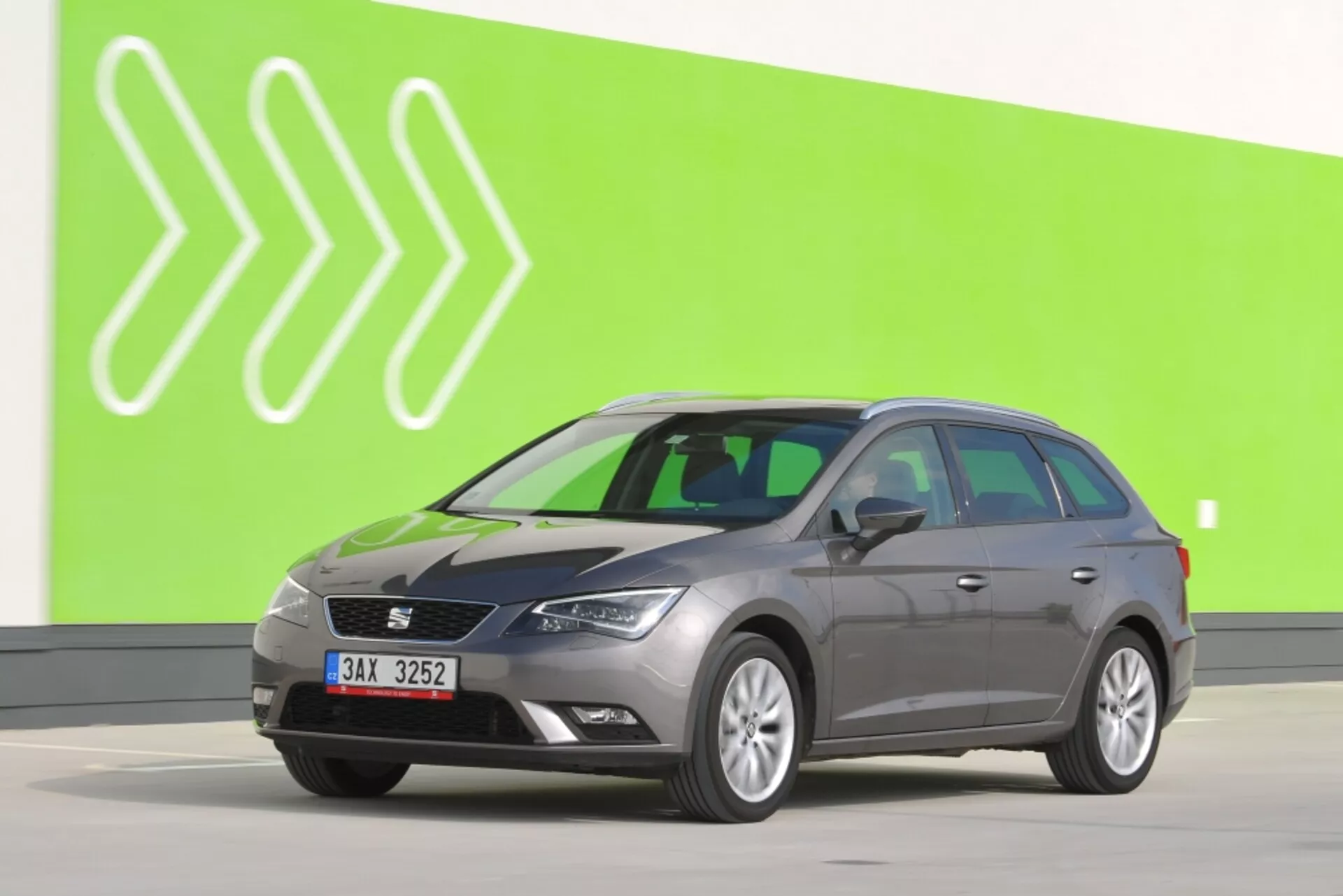 Break gris Seat Leon SC 2012 en vue latérale complète sur fond vert avec flèches blanches stylisées, montrant des rétroviseurs latéraux avec clignotants, des contours de porte caractéristiques et des roues standard sur un sol gris clair de studio