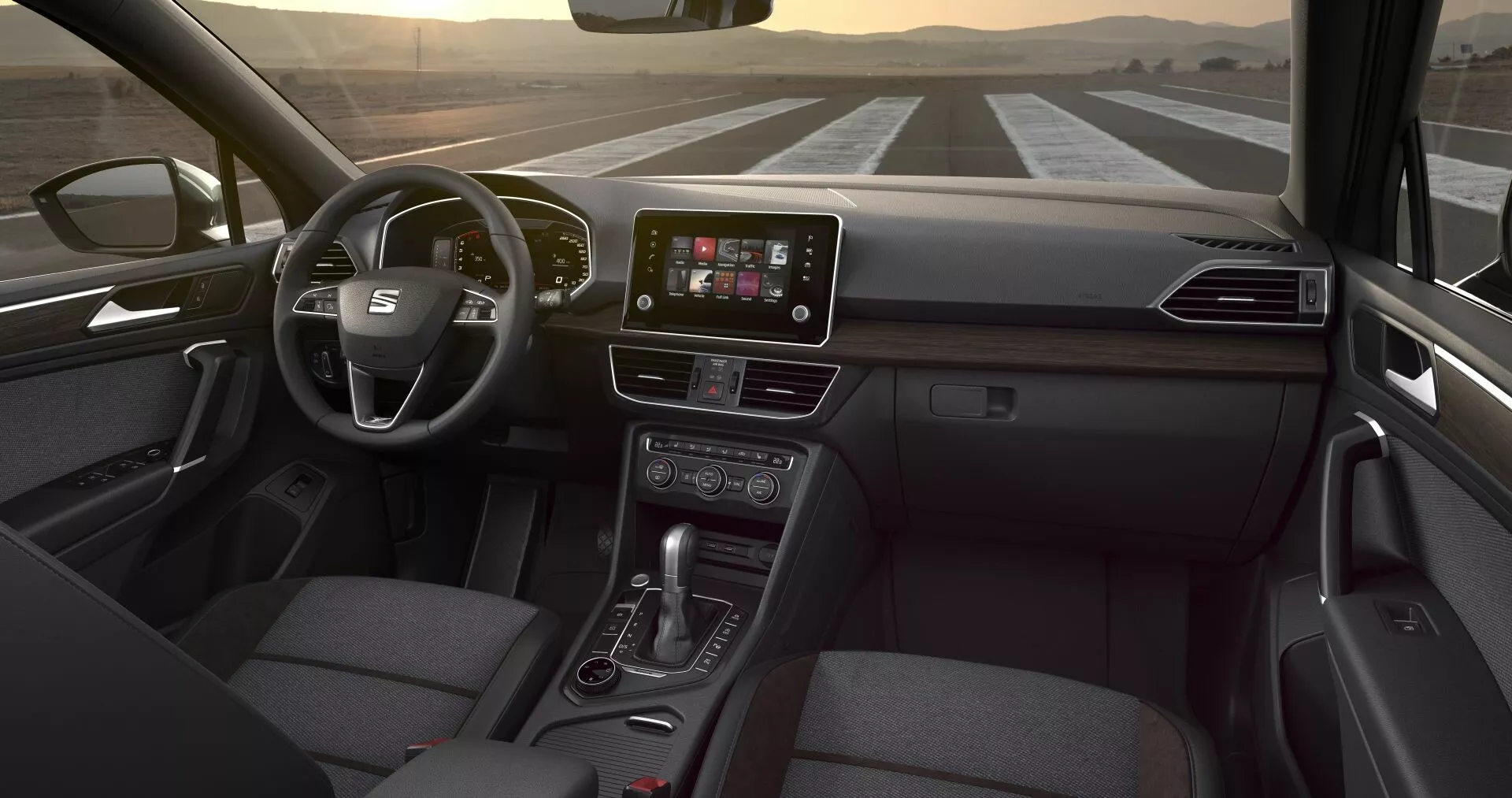 Perspective intérieure du Seat Tarraco 2019 depuis le siège arrière, montrant le volant multifonction, l'écran tactile central pour l'infodivertissement, les boutons de contrôle climatique, le levier de transmission automatique et le siège passager en tissu, sous un éclairage intérieur subtil