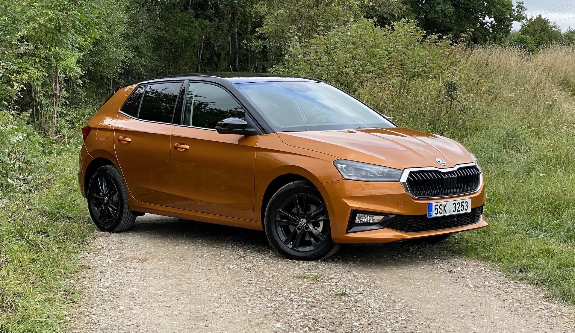 Image d'une Škoda Fabia 2021 moderne de type hatchback, capturée sous un angle frontal trois-quarts, montrant le côté avant-gauche, la porte du conducteur, le capot gauche et une partie du toit. Le véhicule se trouve sur un chemin non pavé entouré de verdure, indiquant un cadre campagnard. Les caractéristiques visibles incluent une calandre horizontale à lames avec un emblème central, des phares LED aigus, une peinture orange métallisée, des rétroviseurs latéraux de la même couleur que la carrosserie, des roues en alliage noir à plusieurs branches avec des pneus à bande de roulement et des portes aux lignes contournées et élégantes. La voiture semble neuve, compacte et non modifiée.