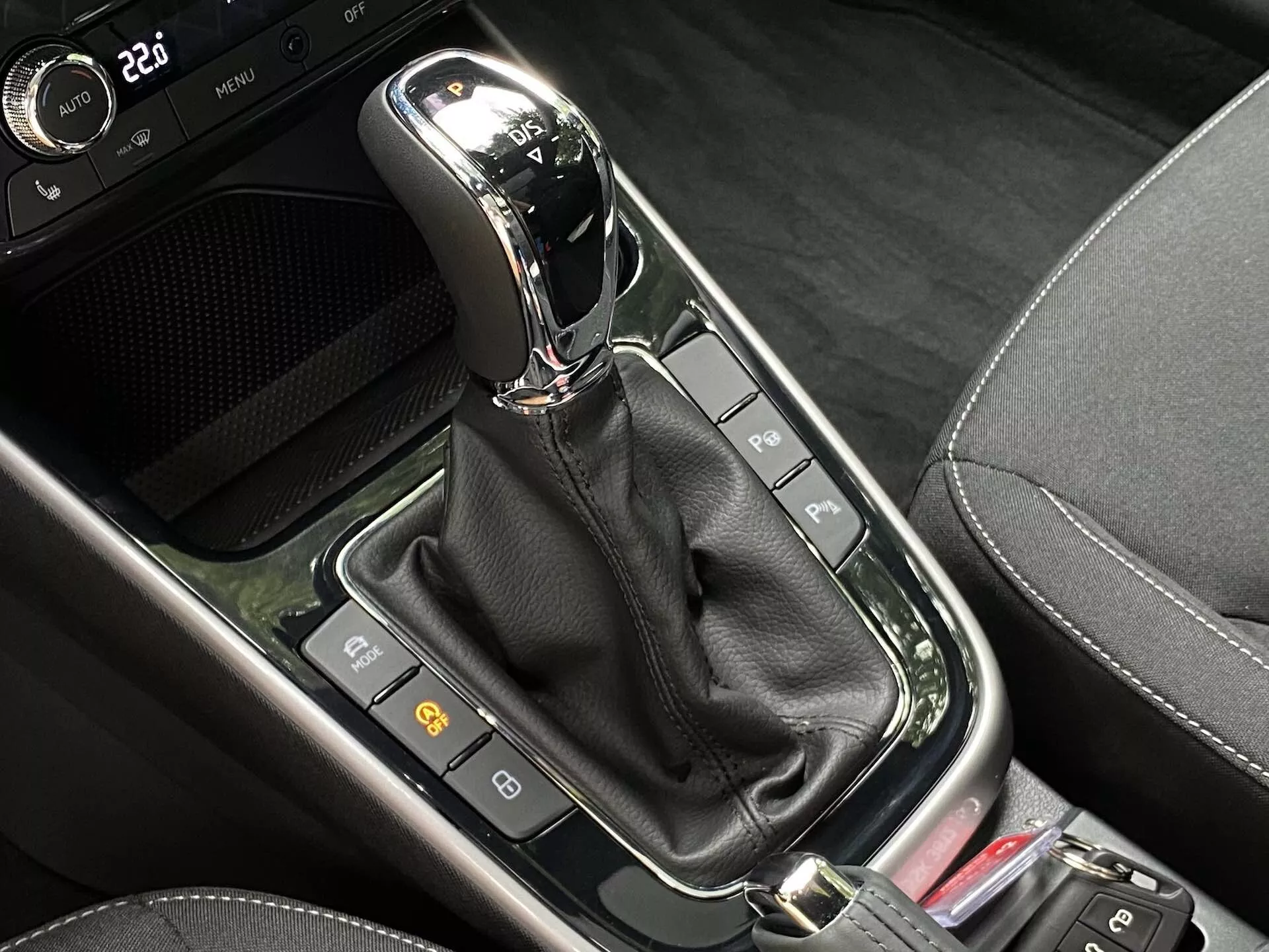 Vue en gros plan du levier de vitesses automatique dans la console centrale d'une Skoda Fabia 2021. Le levier de vitesses moderne, positionné verticalement en mode 'P' (Parking), présente une finition chromée ou métallique sur le dessus et est encapsulé dans un panneau noir brillant. En dessous, une gaine en cuir noir cache le mécanisme. Autour du levier de vitesses, une série de boutons et d'indicateurs, dont deux à gauche probablement liés aux fonctions de sécurité de la voiture, et une partie d'un bouton rouge à droite. La zone mélange divers matériaux noirs, y compris des surfaces brillantes et des motifs imitant la fibre de carbone, suggérant un design contemporain de haute technologie.