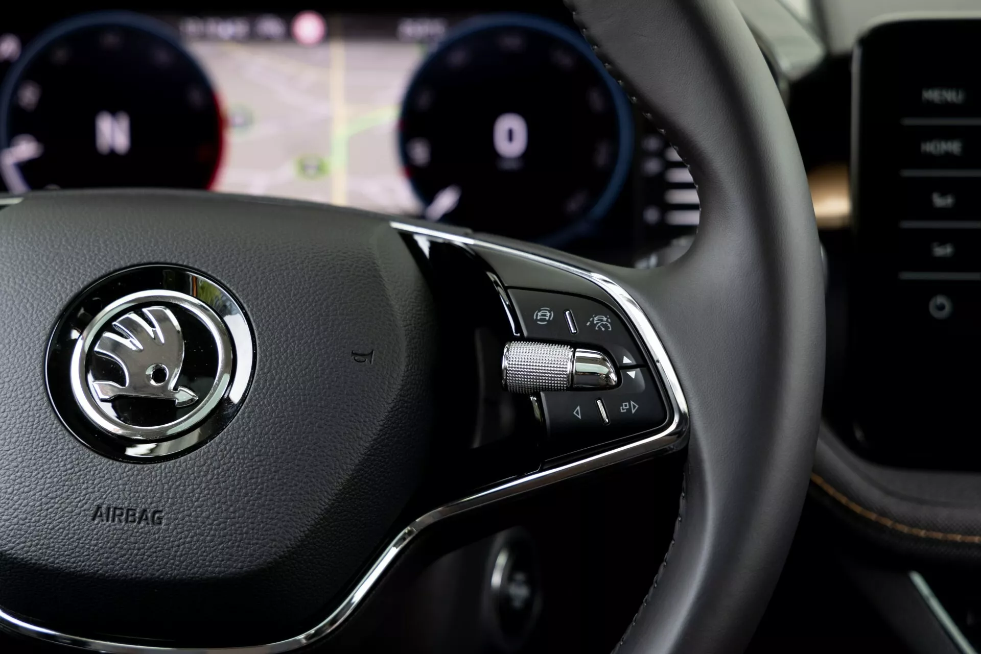 Vue rapprochée de l'intérieur de la Skoda Fabia 2021 depuis la perspective du conducteur, se concentrant sur les commandes du volant à droite. Le volant en cuir présente un emblème argenté central avec le logo Skoda et le texte 'AIRBAG' embossé en dessous. Le groupe de commandes sur la droite inclut des boutons pour le système d'infodivertissement, les appels mains libres ou les commandes vocales, avec des palettes de changement de vitesse visibles derrière le volant. Arrière-plan flou montrant le tableau de bord avec compte-tours et compteur de vitesse, ainsi qu'un écran digital pour l'assistance au conducteur. Console centrale ou système multimédia partiellement visible à droite.