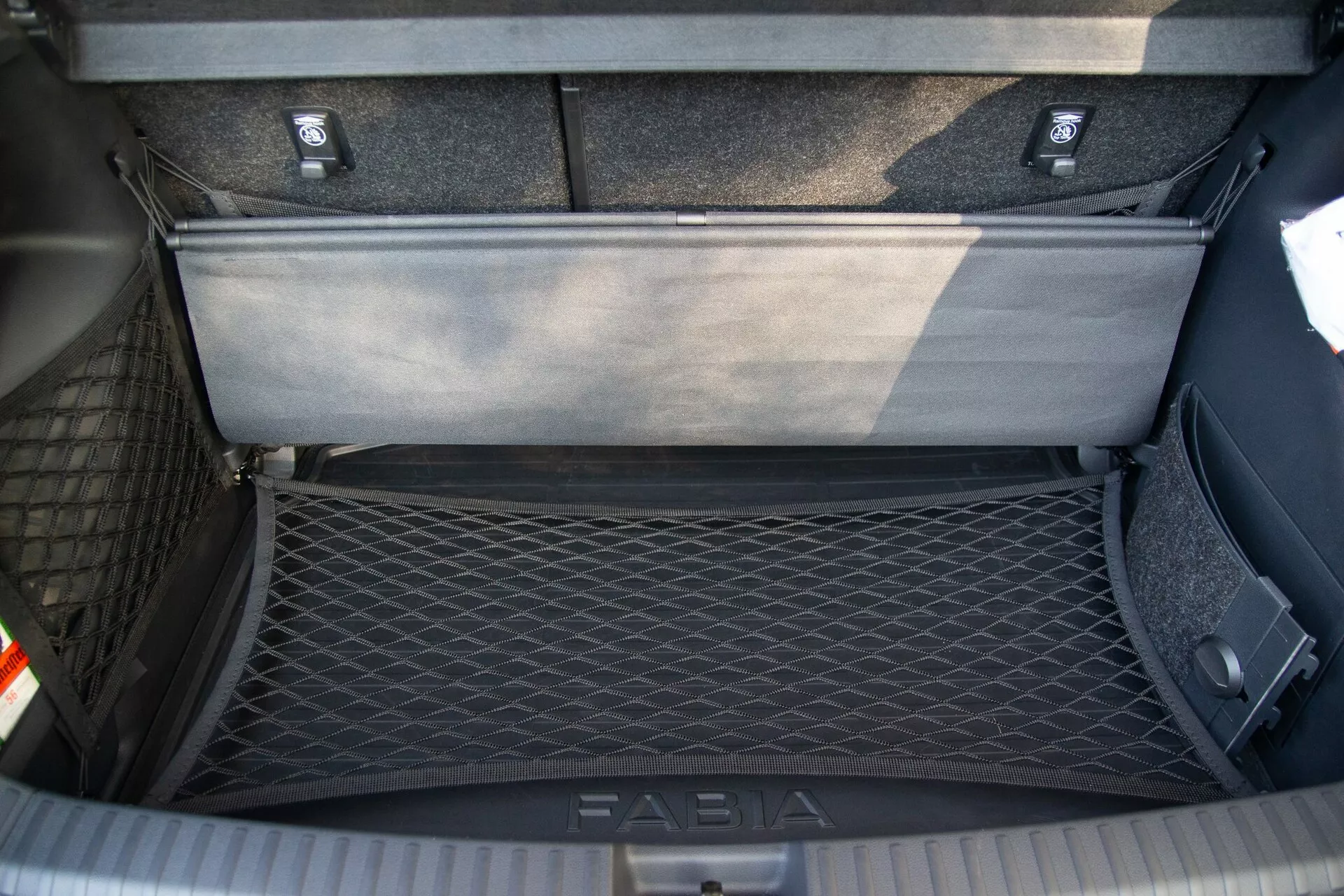 Vue de dessus de la zone de coffre d'une Skoda Fabia 2021, montrant le compartiment de rangement intérieur détaillé avec un filet de chargement sur le côté gauche pour la rétention des objets et un panneau de plancher flexible partiellement relevé révélant une zone de rangement supplémentaire en dessous. Le coffre dispose de moquettes durables et d'une plaque de seuil portant la marque 'FABIA' pour la protection pendant le chargement et le déchargement.