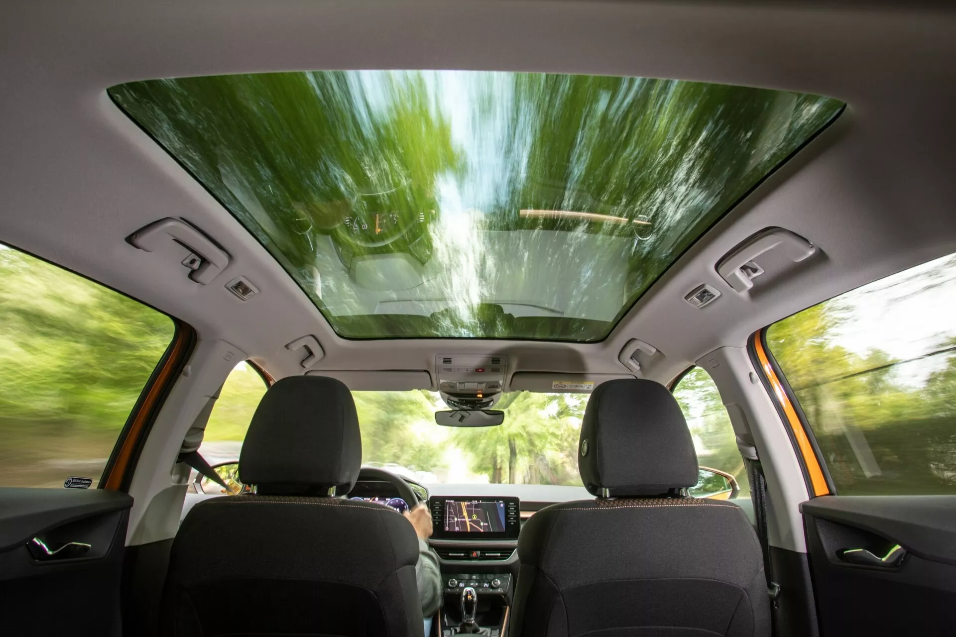 Vue intérieure de la Skoda Fabia 2021 depuis la perspective du siège arrière, montrant les sièges avant avec rembourrage sombre, toit ouvrant panoramique, écran tactile d'infodivertissement allumé sur le tableau de bord, panneaux de commande, haut-parleurs intégrés dans les portes et commandes des vitres, rétroviseur avec des fonctionnalités high-tech possibles et une console supérieure pour des commandes supplémentaires, suggérant une expérience de conduite moderne, confortable et améliorée technologiquement.