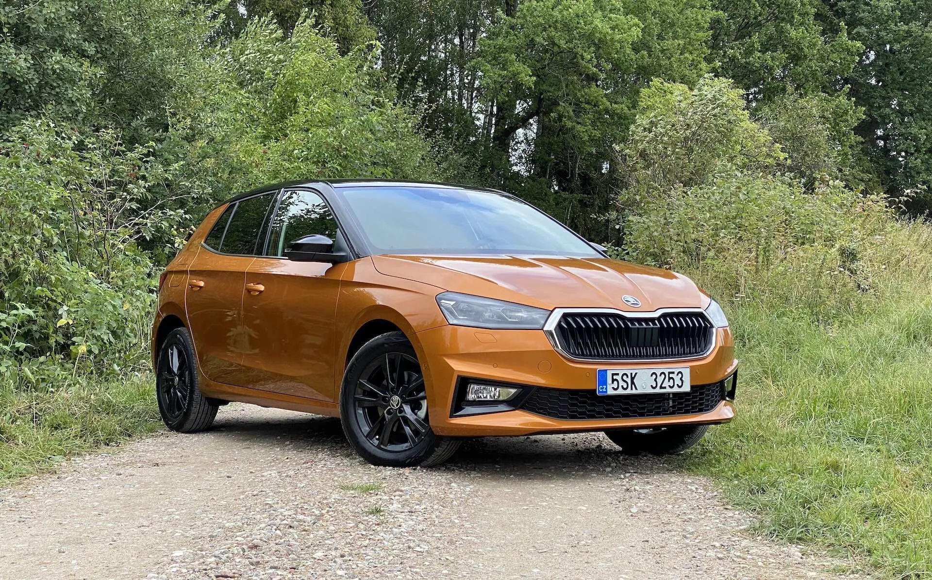 Profil latéral complet d'une Skoda Fabia 2021 moderne de couleur orange garée sur un chemin de terre avec de la végétation en arrière-plan, capturé sous un angle légèrement oblique depuis l'avant, mettant en évidence le côté conducteur. Les caractéristiques incluent une calandre proéminente avec l'emblème de la marque centré, des phares élégants, des phares antibrouillard, de grandes roues en alliage noires à plusieurs rayons, des poignées de porte et des rétroviseurs de la couleur de la carrosserie, une ligne de toit inclinée et un aperçu du hayon arrière et de l'ensemble des feux.