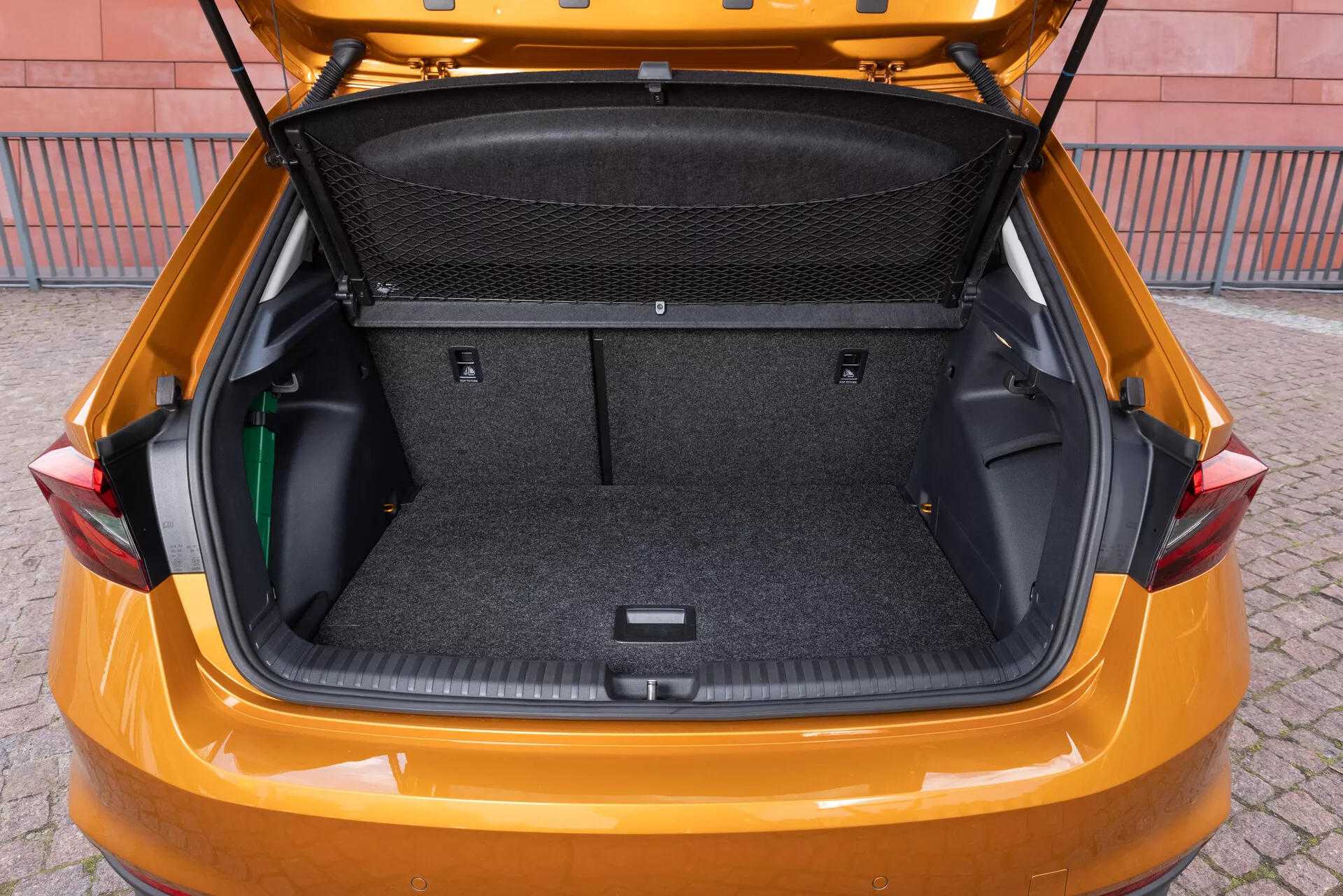L'image représente le coffre d'une Skoda Fabia modèle 2021, prise sous un angle arrière légèrement élevé pour montrer l'espace de chargement spacieux. L'intérieur est revêtu d'un matériau semblable à du tapis et dispose de zones de rangement latérales flexibles, d'une cloison en filet pour la charge et d'un espace encastré au fond pour un rangement supplémentaire. Le hayon est levé, révélant un grand coffre propre, conçu pour mettre en valeur la capacité de rangement et la praticité du véhicule.
