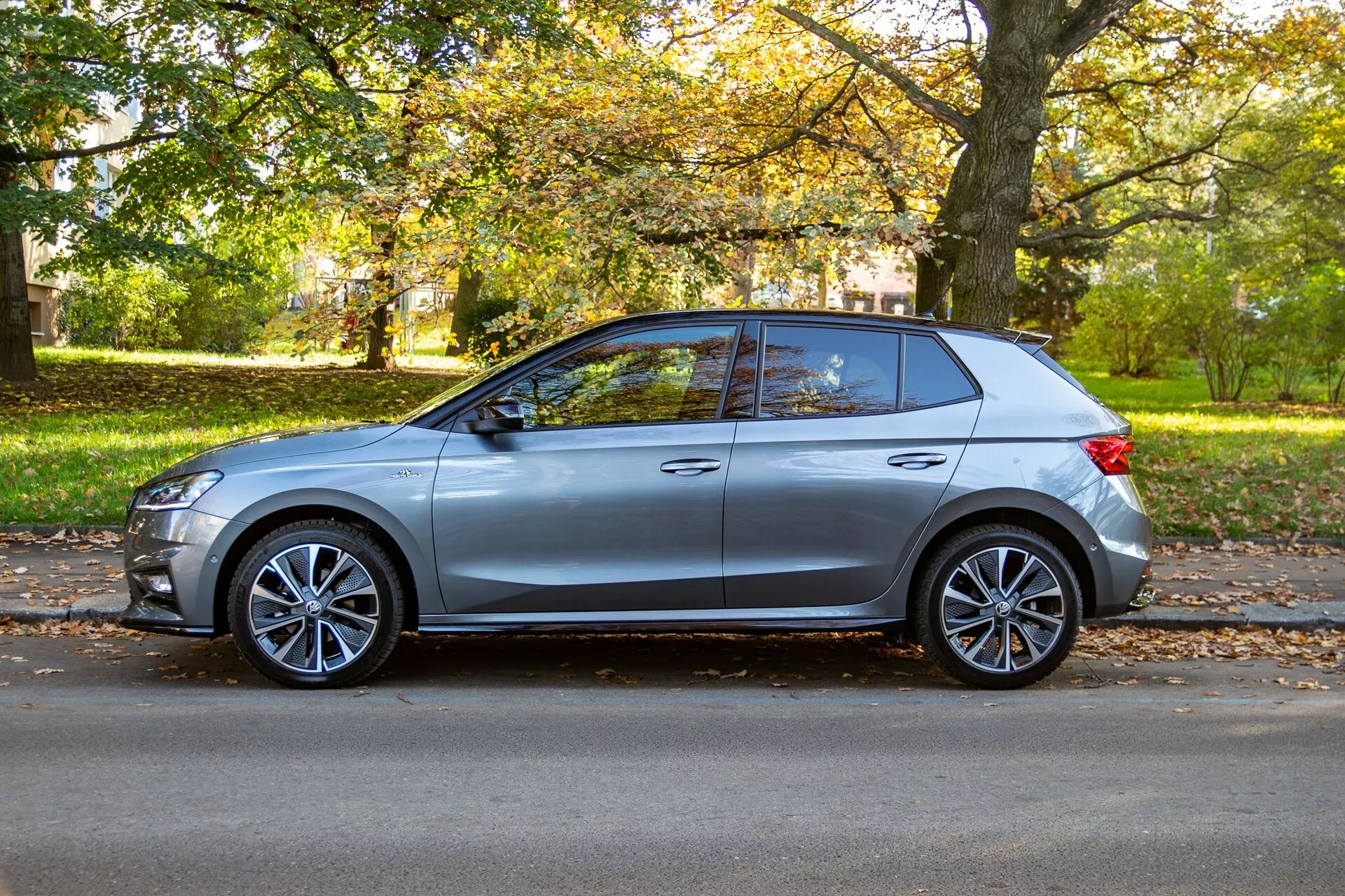 Vue de profil d'une Skoda Fabia 2021 argentée hatchback, garée à côté d'une route bordée d'arbres en plein jour, mettant en avant son design moderne avec une ligne de ceinture haute, des jantes sport en alliage, des poignées de portes affleurantes, des rétroviseurs latéraux aérodynamiques avec clignotants intégrés, une ligne de toit inclinée, des lignes de carrosserie nettes, des vitres partiellement teintées et un montant B noir. L'environnement serein suburbain ou de parc accentue l'esthétique du véhicule.