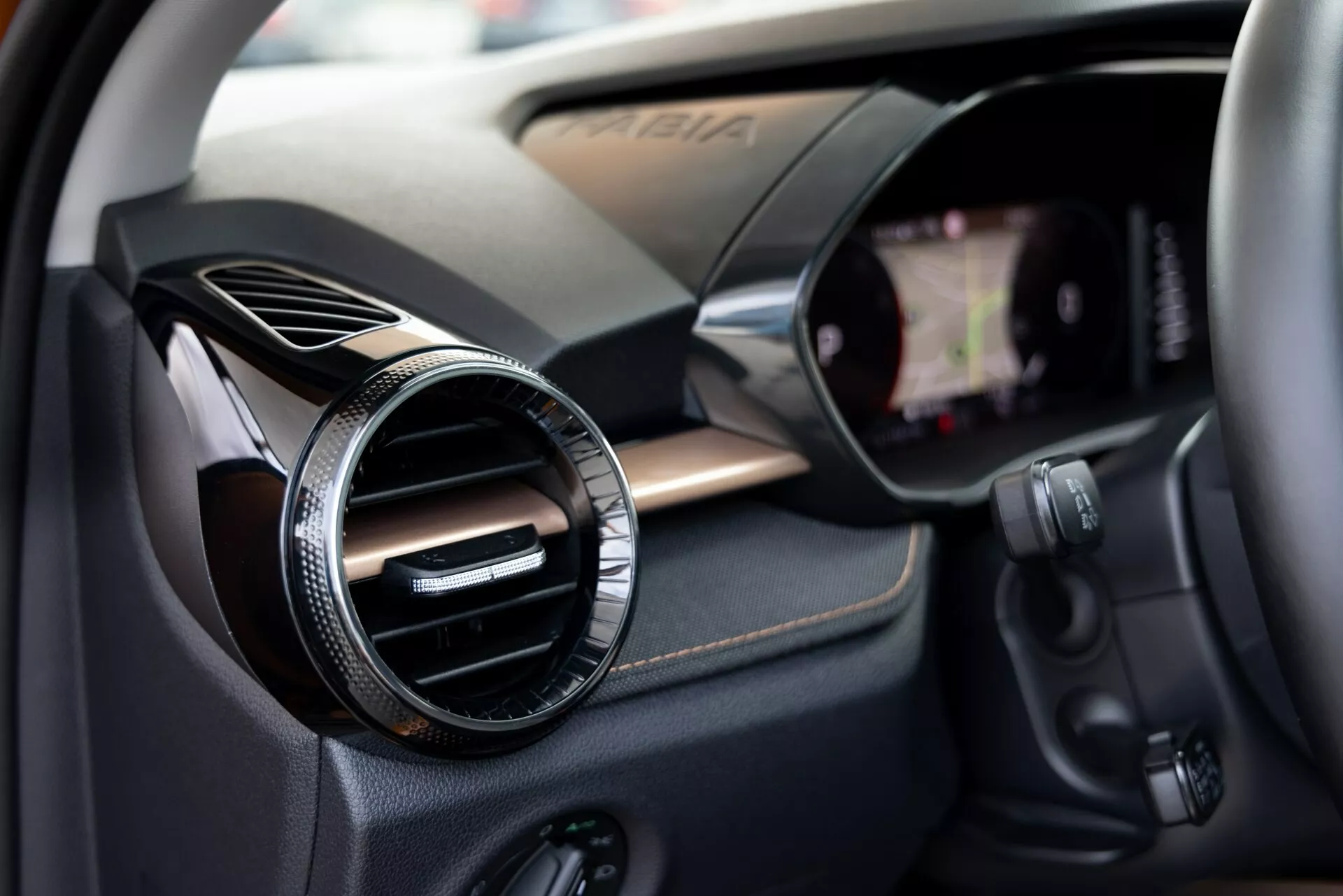 Vue rapprochée de l'intérieur de la Škoda Fabia 2021 axée sur le côté passager. L'image capture une sortie d'air ronde avec une finition métallique ou chromée contre un panneau texturé en fibre de carbone, suggérant une finition sportive ou haut de gamme. À droite, le bord d'un écran tactile d'infodivertissement intégré au tableau de bord est visible. Sous la sortie d'air, on trouve divers boutons de commande, probablement pour le système audio, la climatisation ou d'autres fonctions du véhicule. La faible profondeur de champ attire l'attention sur la sortie d'air tout en suggérant doucement les composants technologiques environnants.