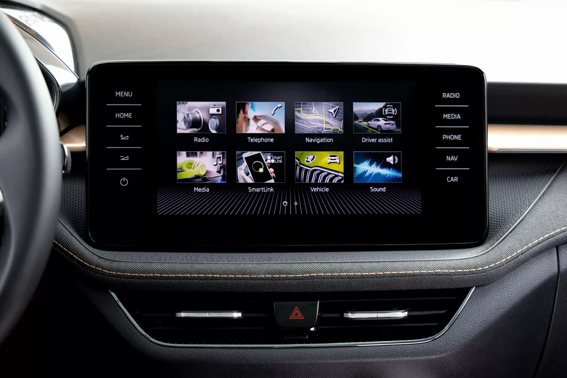 Vue rapprochée du système infotainment à écran tactile de la Škoda Fabia 2021 depuis la perspective d'un passager ou conducteur avant, montrant une console centrale élégante encadrée en ce qui semble être de l'aluminium brossé. L'écran affiche une interface graphique conviviale avec des icônes colorées pour Maison, Radio, Téléphone, Navigation, et plus encore. Sous l'écran, une ligne de boutons physiques et une possible fente pour CD ajoutent des fonctionnalités multiples. La sellerie haut de gamme avec des surpiqûres contrastées suggère un design intérieur de haute qualité.