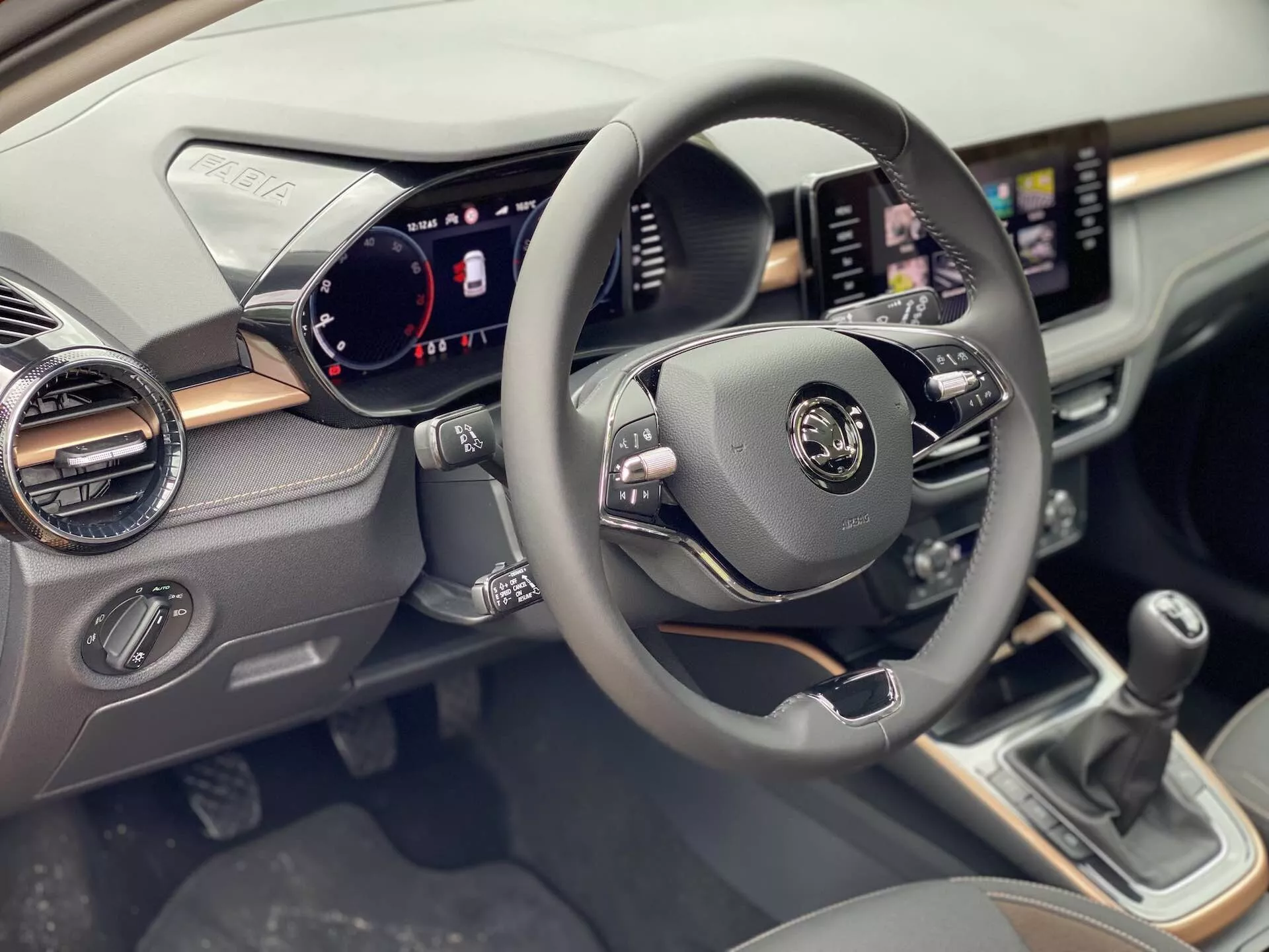 Vue intérieure de la Skoda Fabia 2021 centrée sur le côté conducteur, montrant le volant au centre avec des commandes multifonctionnelles pour l'audio, les commandes vocales et le régulateur de vitesse. Le tableau de bord numérique affiche le compteur de vitesse, le compte-tours et les indicateurs de statut du véhicule. Un système d'infodivertissement à écran tactile est visible sur le tableau de bord avec des fonctionnalités de navigation et de médias. Sous l'écran se trouvent des cadrans et des boutons de contrôle climatique. Le levier de changement de vitesse manuel est visible sur la console centrale.
