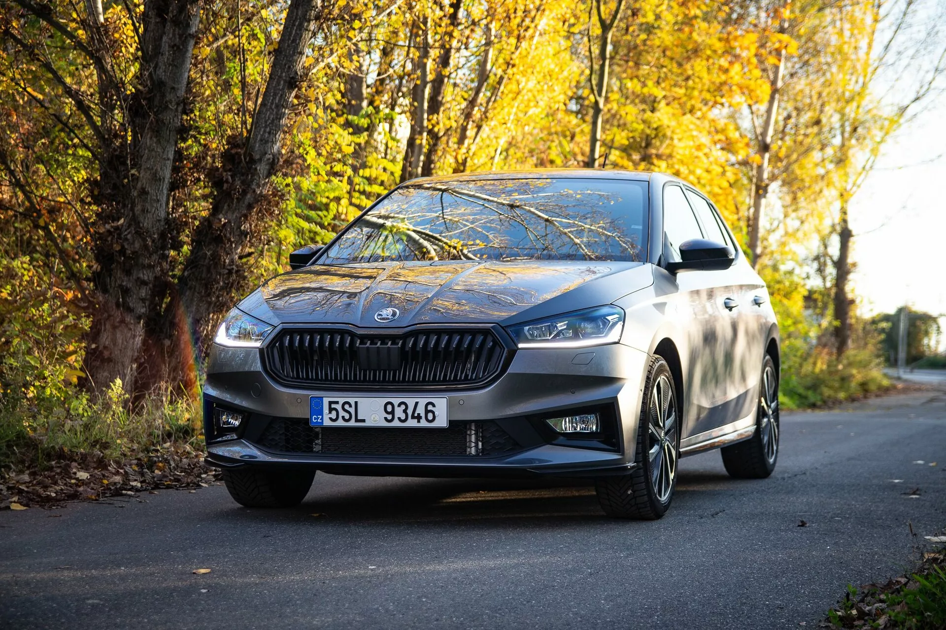 Image d'une berline moderne Skoda Fabia 2021 garée sur une route bordée de feuillage automnal, capturée depuis une perspective au niveau du sol, mettant en avant la calandre avant, les phares angulaires, les lignes de design élégantes, les roues en alliage et les rétroviseurs latéraux avec clignotants intégrés, reflétant une finition métallique de haute qualité et une combinaison de luxe et de sportivité.