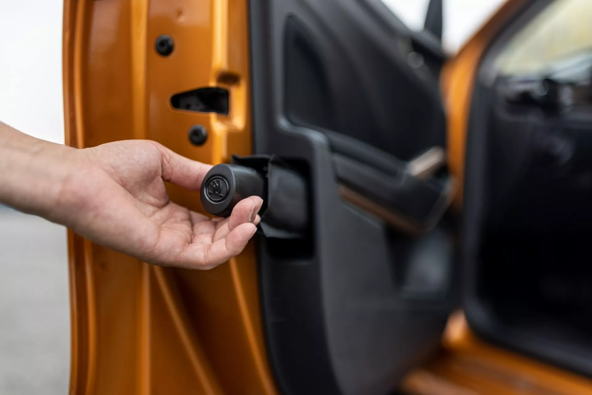 Gros plan sur la porte côté conducteur de la Skoda Fabia 2021, légèrement entrouverte, vue de l'extérieur avec le bord intérieur visible. Une main tient un porte-clés noir et circulaire, compact, indiquant un système d'entrée sans clé ou de sécurité automobile. La voiture est orange vif avec une finition métallique, mettant en valeur des mécanismes de verrouillage avancés et des fonctionnalités de sécurité personnalisées.