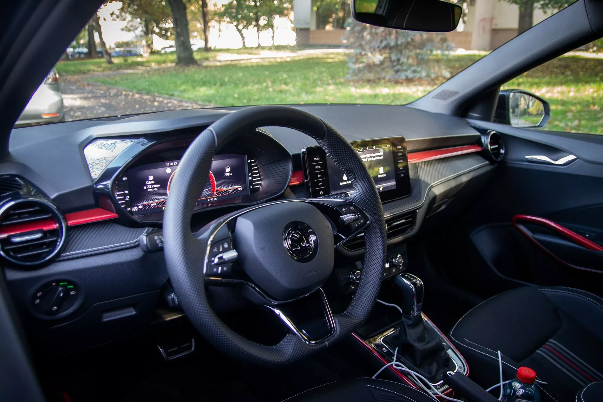 Vue intérieure d'une Skoda Fabia 2021 moderne depuis la perspective du conducteur, avec panneau de contrôle sur la porte du conducteur, volant multifonctionnel, tableau de bord numérique, aérateurs accentués de rouge, écran tactile central d'infodivertissement, levier de vitesse manuel et zone siège passager avec boîte à gants et aérateur assorti.