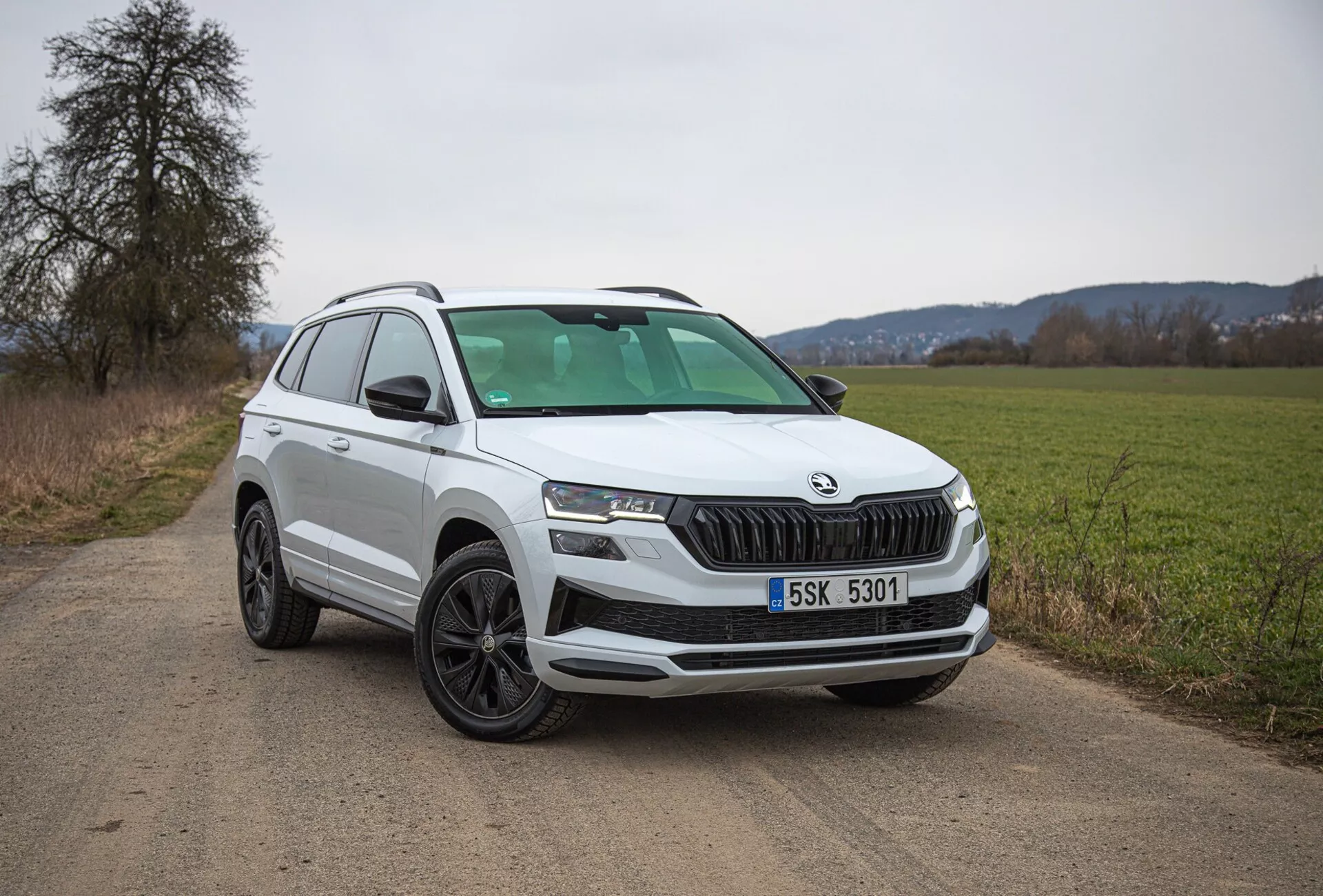 SUV blanc Škoda Karoq 2021 garé sur une route de campagne en terre, présentant la vue du quart avant gauche incluant la grille, les phares et le design des roues, avec un accent sur les caractéristiques modernes telles que l'éclairage LED et les capteurs de stationnement.