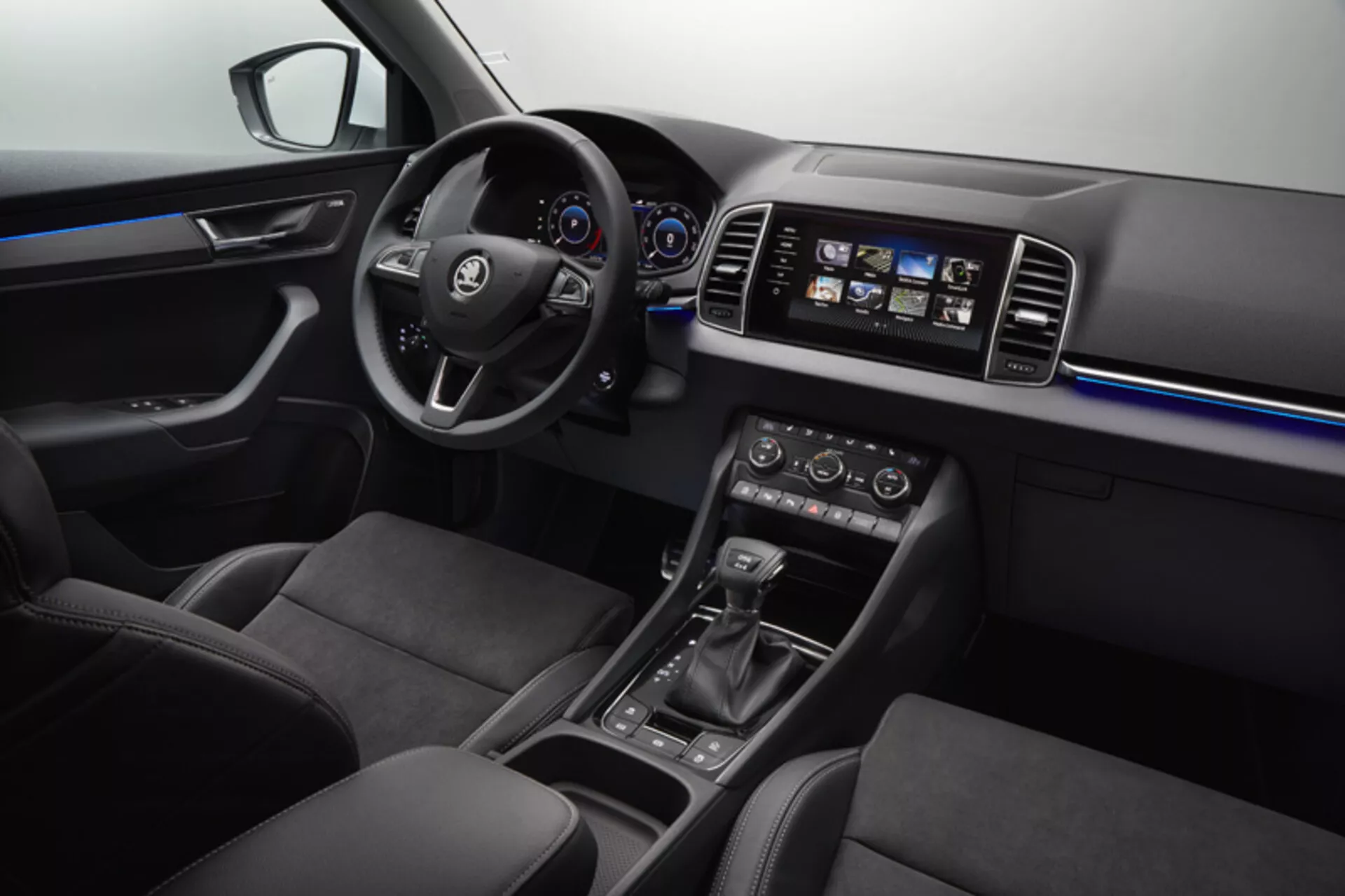 Vue intérieure du Skoda Karoq 2017 montrant le tableau de bord, porte du conducteur avec contrôles des fenêtres et des rétroviseurs, volant avec boutons multimédia et régulateur de vitesse, combiné d'instruments numérique-analogique, système d'infodivertissement tactile, contrôle climatique bi-zone, sélecteur de vitesse automatique, sélection du mode de conduite et sièges en cuir