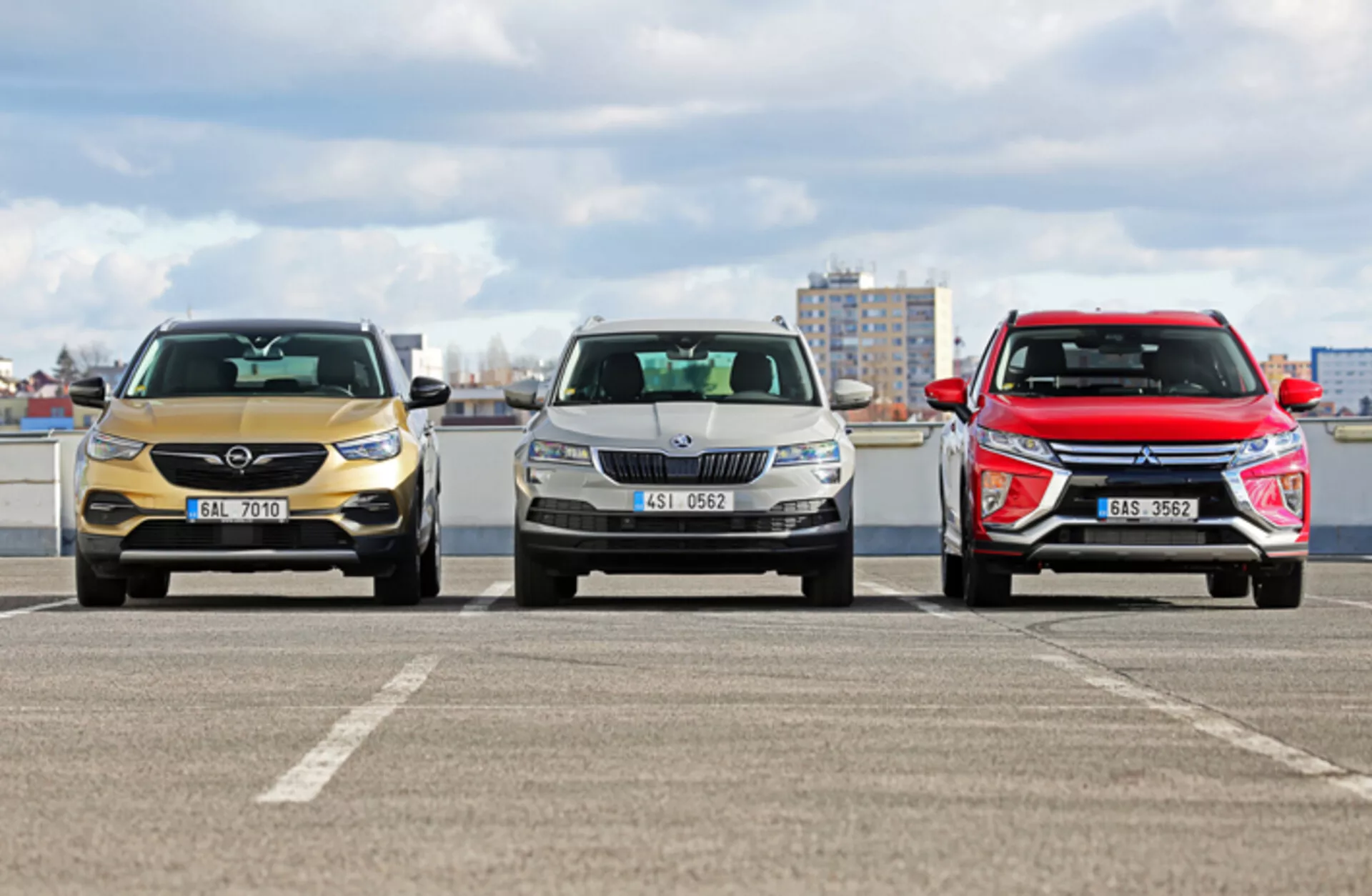 Vue de face de trois modèles de Skoda Karoq 2017 sur un parking avec un arrière-plan de paysage urbain, montrant différents designs de grilles avant, de phares et de formes de capot aérodynamiques sous des conditions de lumière diurne nuageuse