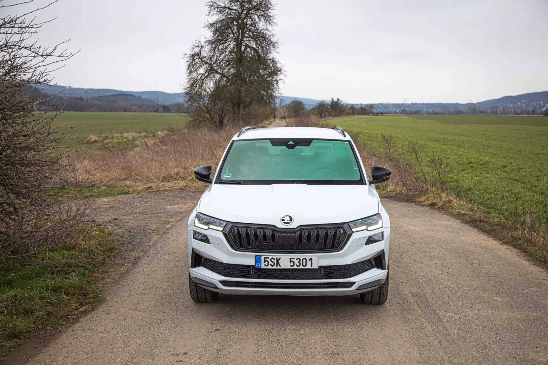 SUV Škoda Karoq 2021 présentant une vue frontale directe sur une route de campagne étroite non pavée, mettant en lumière la grille distinctive de Škoda, des phares LED précis, des feux de brouillard dans un contour noir pour une capacité potentiellement tout-terrain, des lignes dynamiques sur le capot et des fonctionnalités technologiques avancées à l'avant du véhicule.