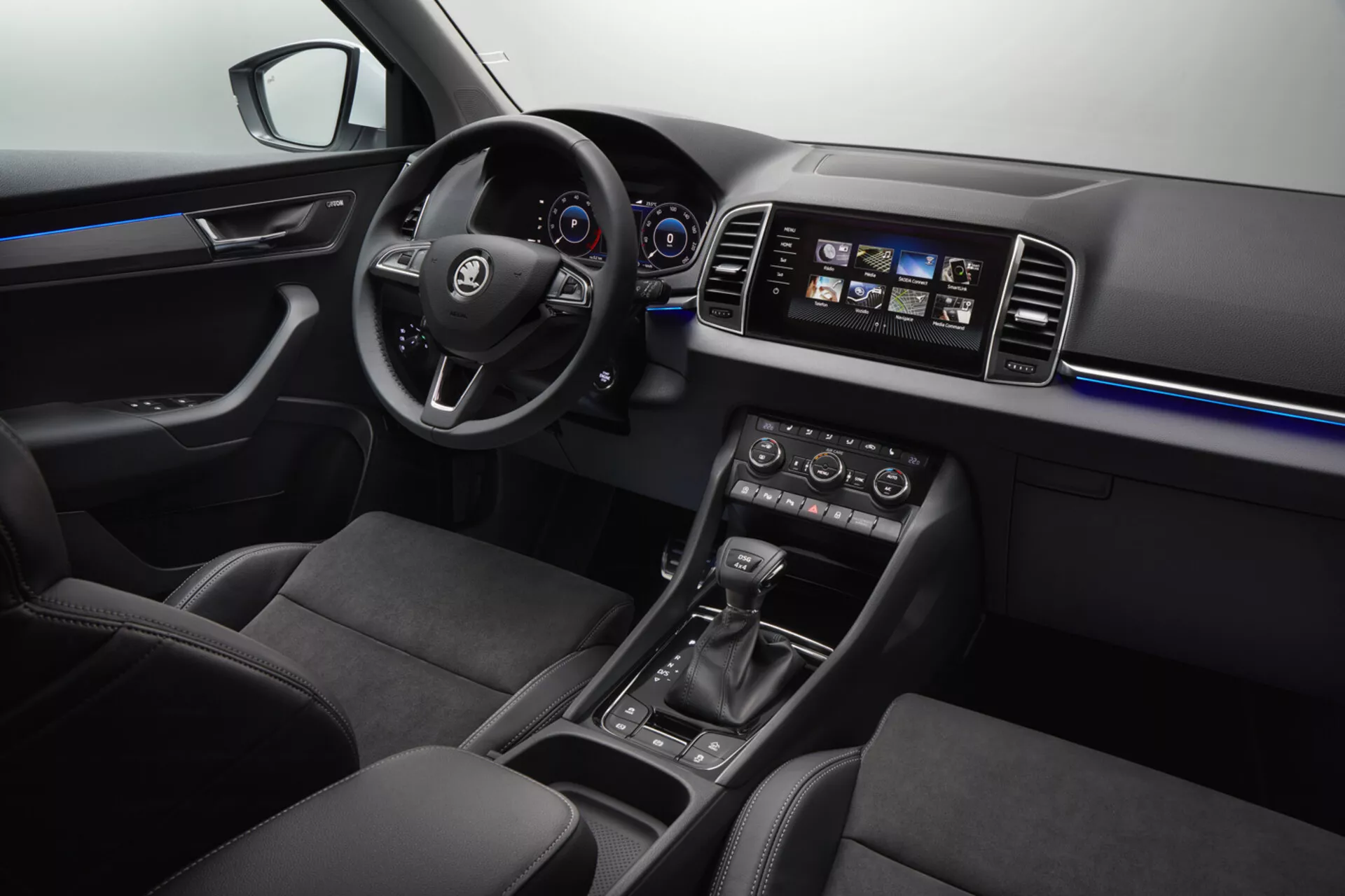 Vue intérieure du Skoda Karoq 2017 mettant en évidence le côté conducteur avec le volant présentant le logo, tableau de bord numérique, console centrale illuminée avec système d'infodivertissement tactile, commande automatique du climat, levier de vitesse avec sélecteurs de mode de conduite, éclairage d'ambiance bleu et sièges en tissu foncé haut de gamme.