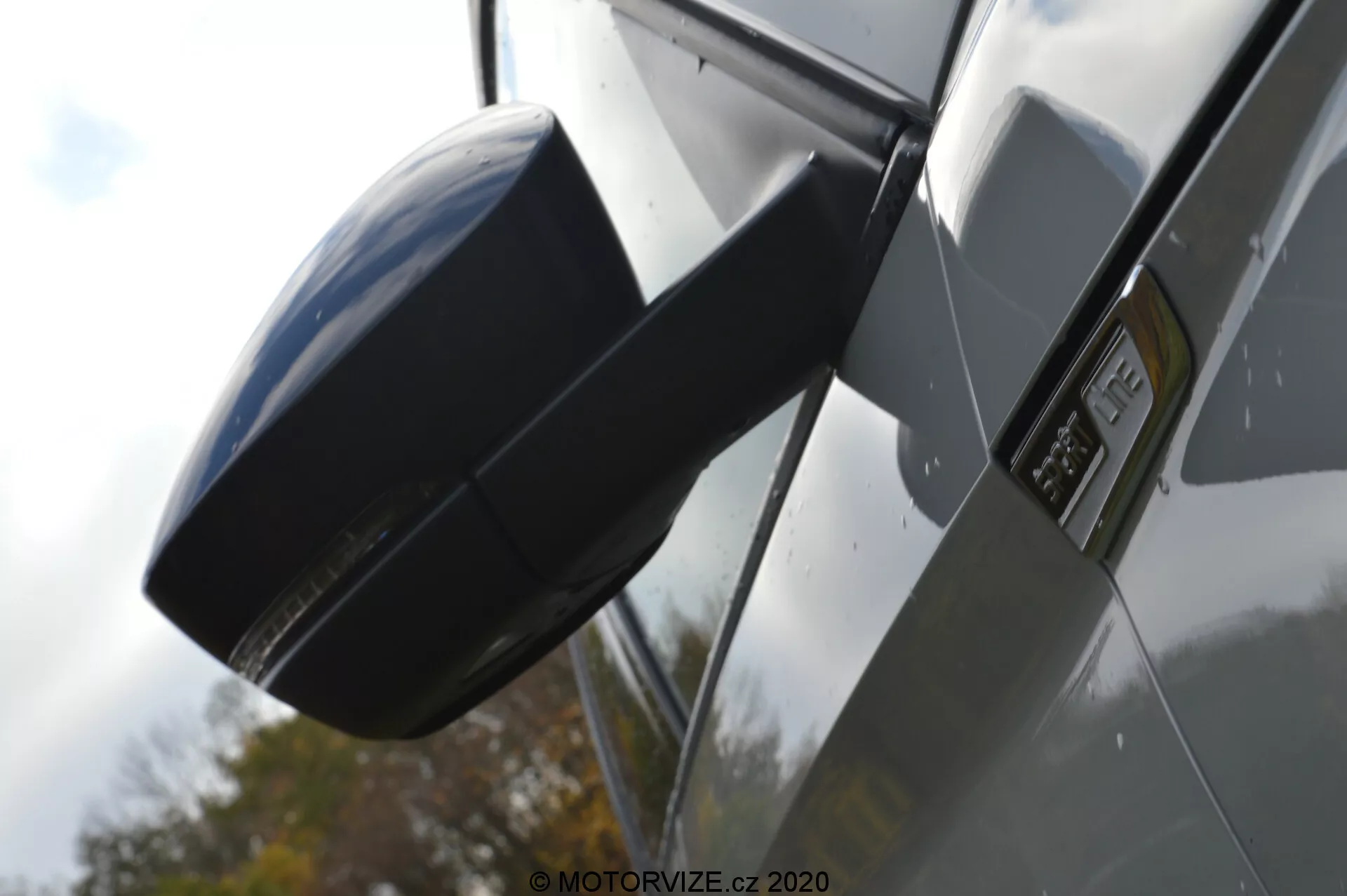 Vue latérale rapprochée d'une Skoda Kodiaq 2017 se concentrant sur le rétroviseur extérieur et la zone du garde-boue. L'image est prise d'un angle bas, mettant en valeur le profil latéral de la voiture, avec un boîtier de rétroviseur latéral moderne et aérodynamique assorti à la couleur du véhicule, un badge proéminent sur le garde-boue indiquant un modèle ou une finition spécifique, des panneaux de carrosserie métalliques et brillants, et des reflets suggérant un environnement extérieur avec une voiture de couleur sombre.