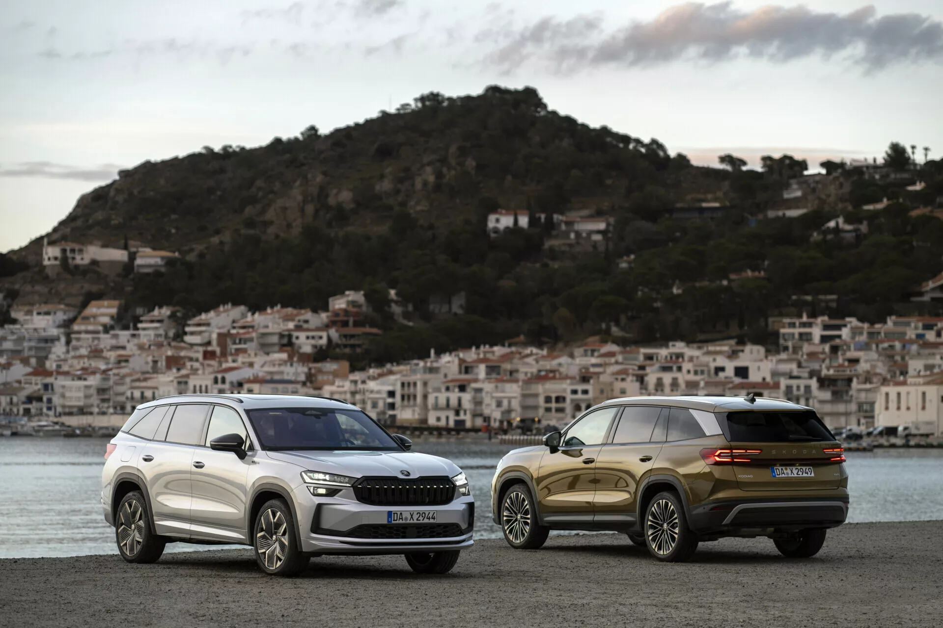 SUV Skoda Kodiaq 2024 garés côte à côte à l'aube sur une promenade en bord de mer avec un village côtier en arrière-plan, montrant la grille avant, les phares et les lignes de design élégantes de l'un et la vue arrière trois quarts de l'autre.