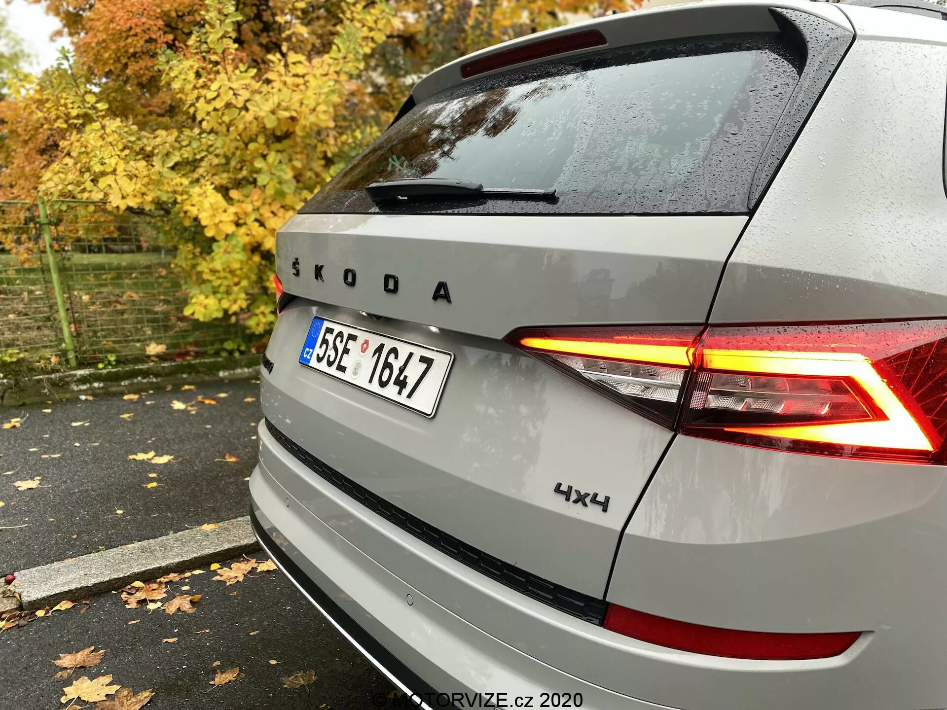 Vue arrière d'un véhicule Škoda argenté avec feux arrière à LED en forme de C illuminés, emblème et marque Škoda, avec badge 4x4, capturée du côté arrière droit sur un trottoir mouillé parmi des feuilles d'automne et des arbres.