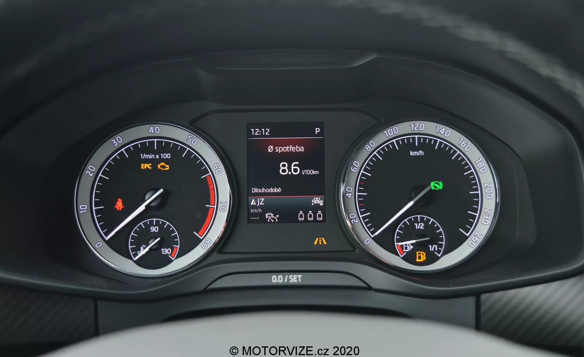 Gros plan depuis la perspective du conducteur sur le tableau de bord de la Skoda Kodiaq 2017, montrant une combinaison de jauges analogiques et numériques derrière le volant. Le tableau de bord comprend un compte-tours avec un voyant orange, un écran central affichant diverses informations sur le véhicule telles que l'heure, l'état du frein de stationnement, le mode sport, l'autonomie et la température, et un compteur de vitesse avec jauge de carburant et indicateur de démarrage/arrêt automatique.
