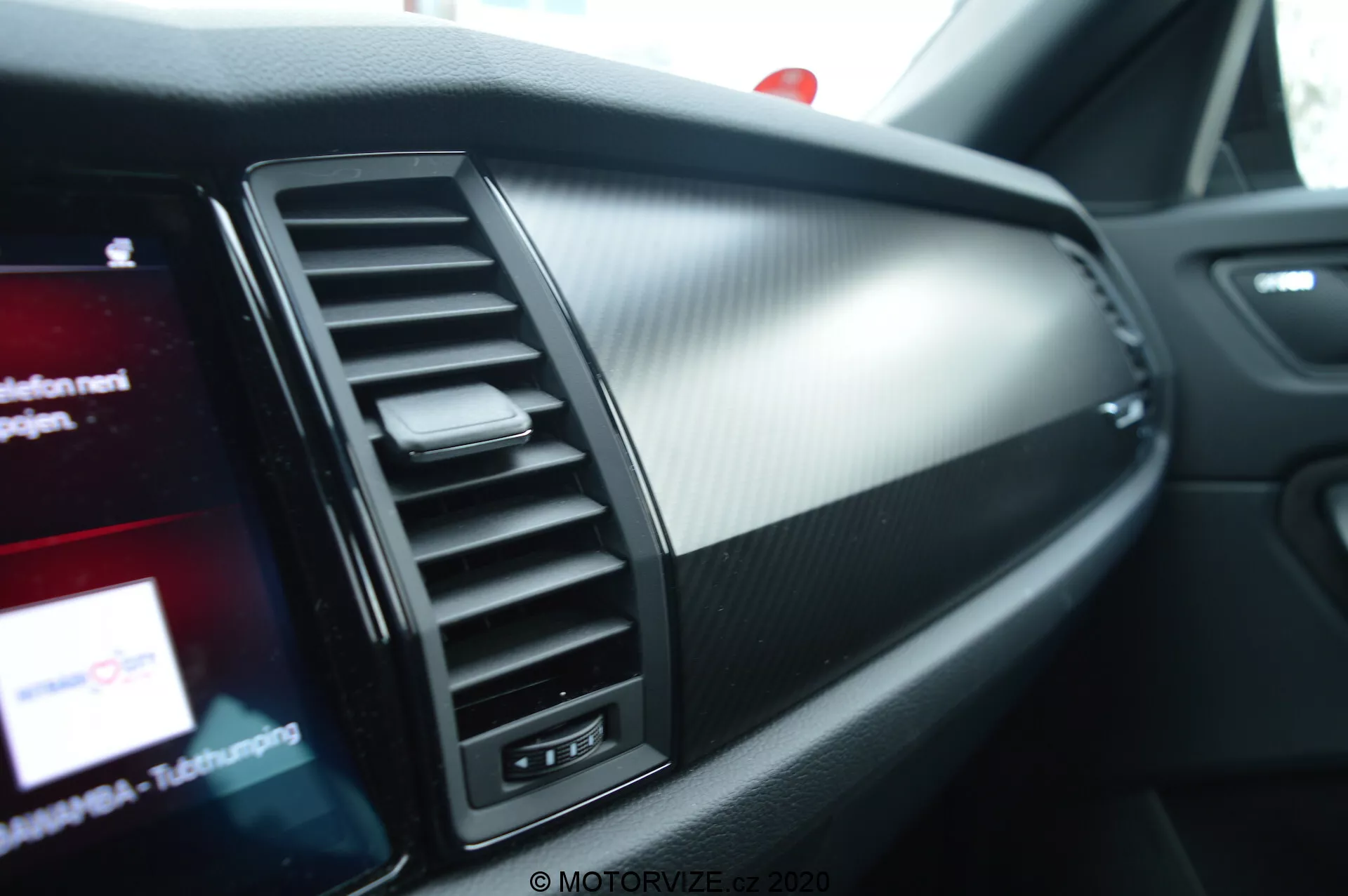 Vue en gros plan de l'intérieur de la Skoda Kodiaq 2017, mettant en avant le côté passager du tableau de bord. Les caractéristiques comprennent une ventilation avec des persiennes réglables verticalement, une section de garniture décorative et un écran d'infodivertissement partiellement visible avec des graphiques colorés. Les matériaux du tableau de bord combinent du métal brossé et du plastique texturé ressemblant à de la fibre de carbone, soulignant un design moderne et sportif.