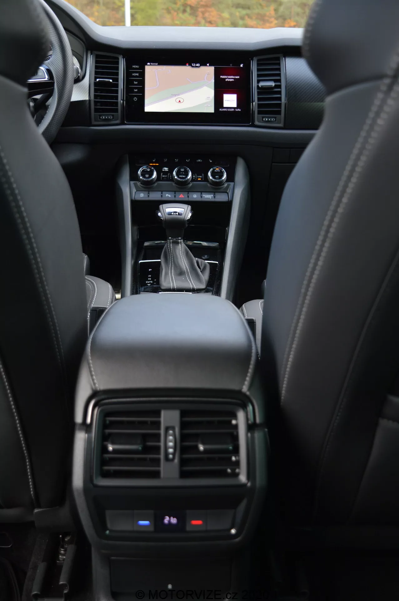 Intérieur de la Skoda Kodiaq 2017, montrant le tableau de bord avant vu depuis la perspective du siège arrière. L'image met en avant les sièges en cuir, la console centrale avec levier de vitesses automatique, les boutons de commande, le système d'infodivertissement à écran tactile affichant une carte de navigation, les bouches d'aération du système de climatisation et une vue partielle du tableau de bord affichant des informations opérationnelles critiques.