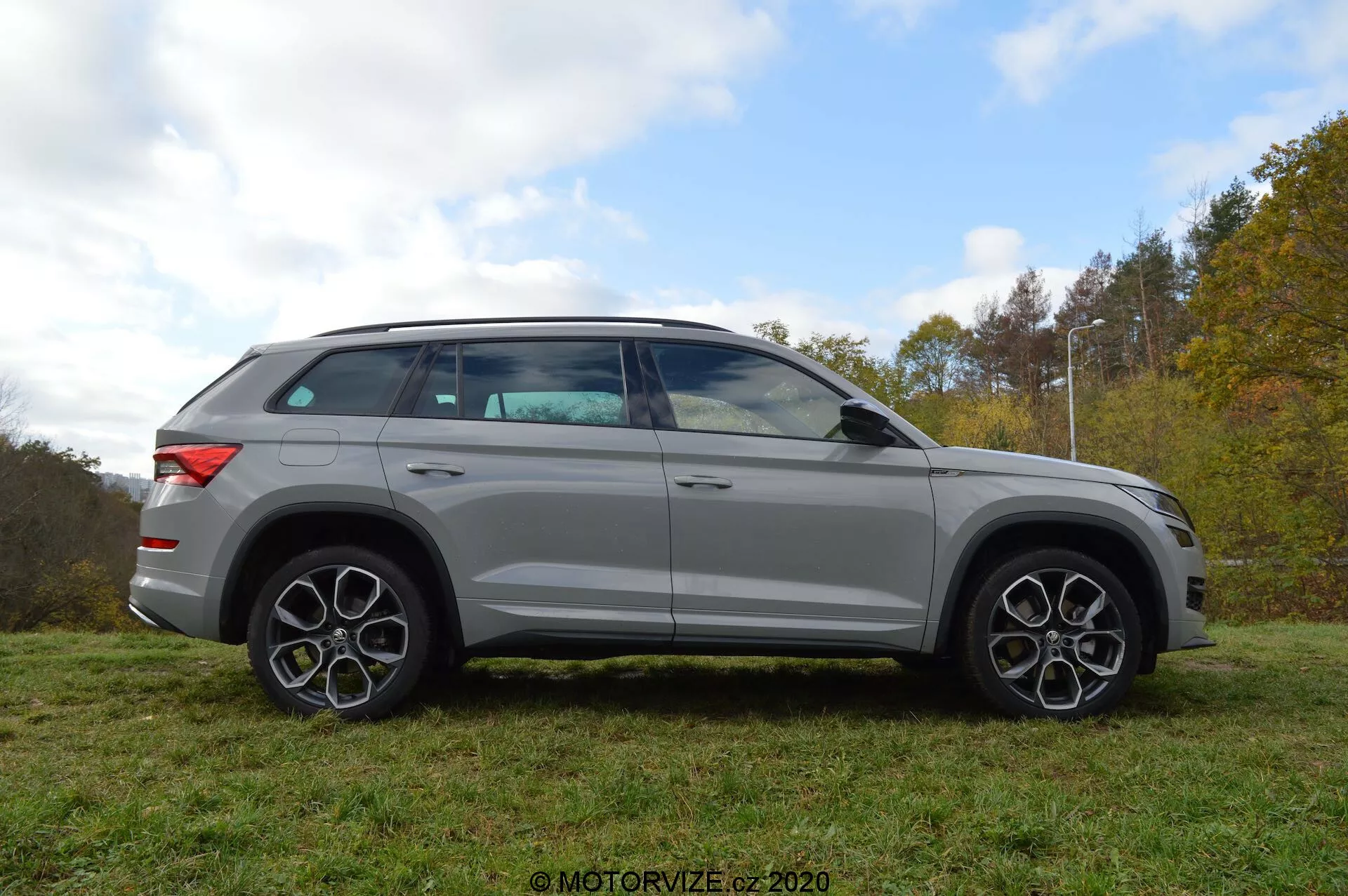 Vue de profil complet d'un SUV Skoda Kodiaq 2017 argenté garé sur l'herbe avec en arrière-plan des arbres et un ciel nuageux. Le design du véhicule se caractérise par des lignes audacieuses, de grandes jantes en alliage et des jantes distinctives à plusieurs rayons. Les rétroviseurs, qui intègrent des clignotants, sont montés juste en dessous de la ligne de fenêtre. Les vitres sont légèrement teintées et la ligne du toit s'incline doucement vers l'arrière, indiquant un profil sportif, semblable à un coupé. Le SUV est également équipé d'un système de rails de toit.