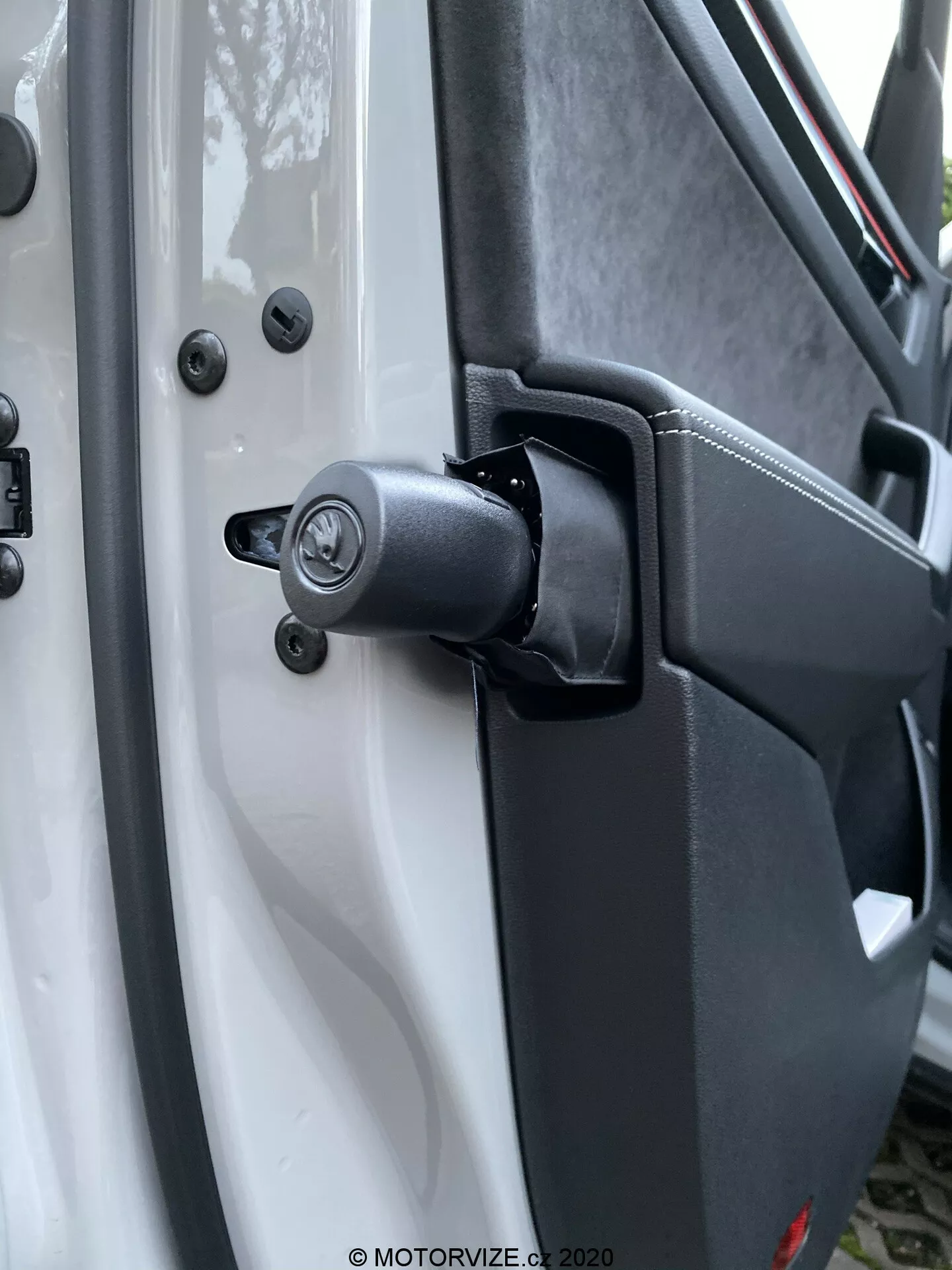 Vue rapprochée du mécanisme de charnière de porte robuste et de la zone environnante sur une Škoda Kodiaq 2017, avec un composant à ressort visible pour une utilisation intensive. Le véhicule blanc contraste avec les composants de charnière noirs, y compris un mécanisme de verrouillage traditionnel avec trou de serrure au-dessus de la charnière, fixé avec des boulons à tête hexagonale pour la durabilité. Le plastique texturé du panneau intérieur de la porte et les points de montage sécurisés renforcent le design utilitaire. L'arrière-plan flou met en évidence la charnière et le mécanisme de verrouillage.