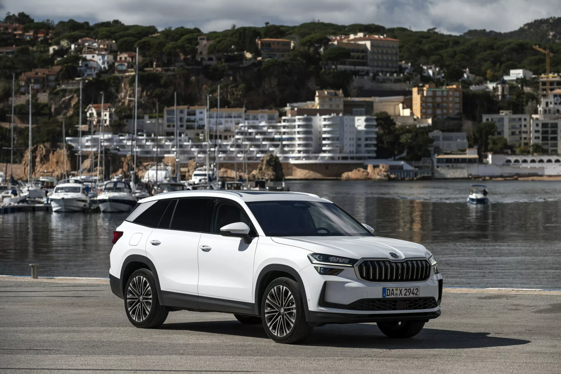 SUV crossover Skoda Kodiaq 2024 garé sur une promenade en bord de mer montrant la vue complète de la voiture incluant des éléments de design moderne comme les phares LED, la grande calandre, les roues en alliage à multiples branches, avec un fond pittoresque de bateaux et de paysage urbain