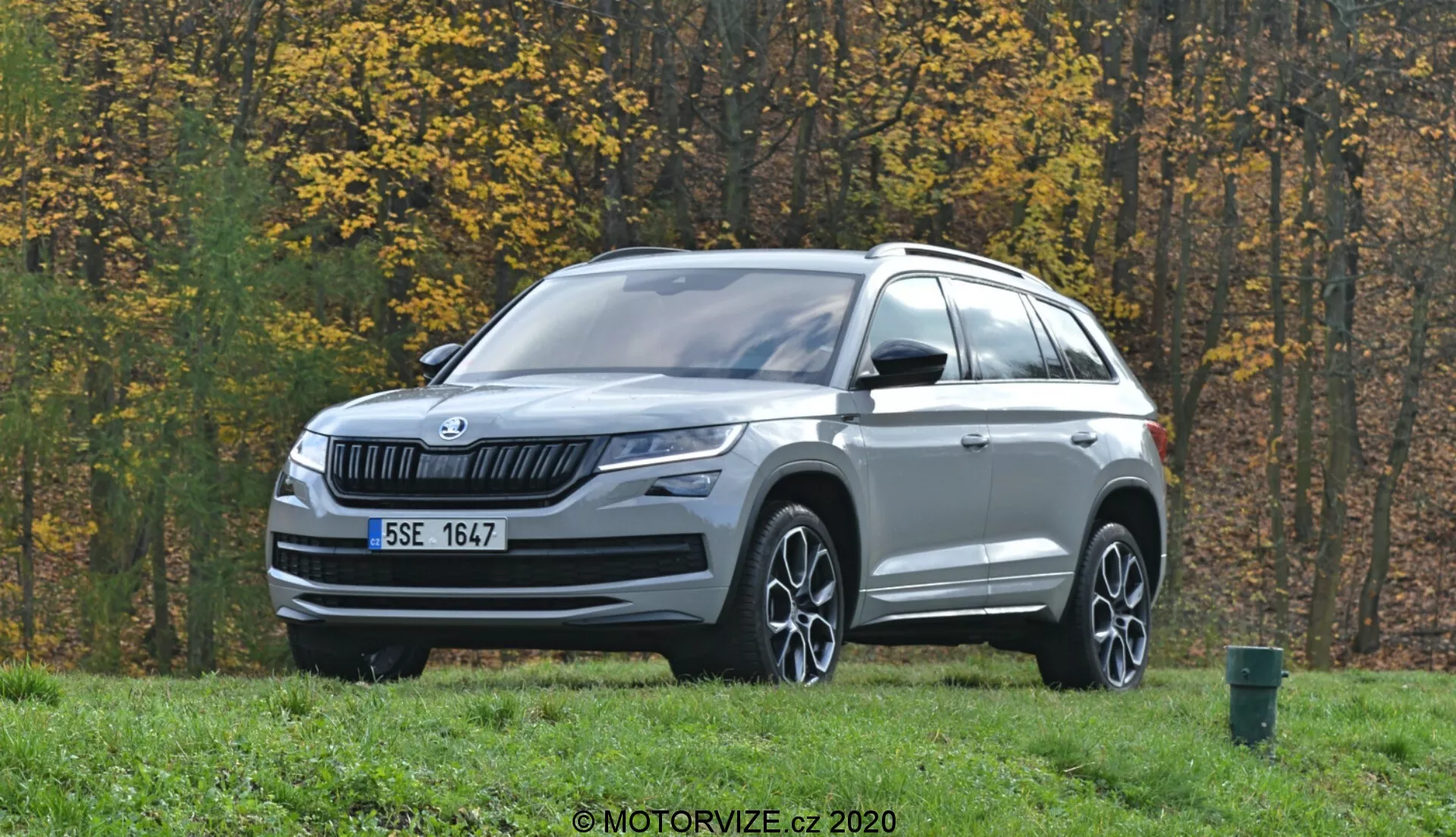 L'image montre un SUV Skoda Kodiaq 2017 garé sur une zone herbeuse avec des arbres d'automne en arrière-plan. Photographié sous un angle de trois quarts de face, le véhicule présente une calandre à lamelles horizontales avec le logo Skoda au centre. Les phares LED élégants s'étendent sur les côtés, accompagnés de feux antibrouillard sur le pare-chocs inférieur. Le capot présente des lignes subtiles et une garniture argentée orne la partie inférieure du pare-chocs avant. Les roues en alliage à rayons multiples, les rétroviseurs avec clignotants intégrés, les barres de toit et les lignes sculptées du corps mettent en valeur le profil. Les capteurs ou caméras sur la partie inférieure du pare-chocs suggèrent des systèmes avancés d'assistance à la conduite (ADAS). Le décor extérieur comprend une route goudronnée et une poubelle verte sur le bord droit de l'image.