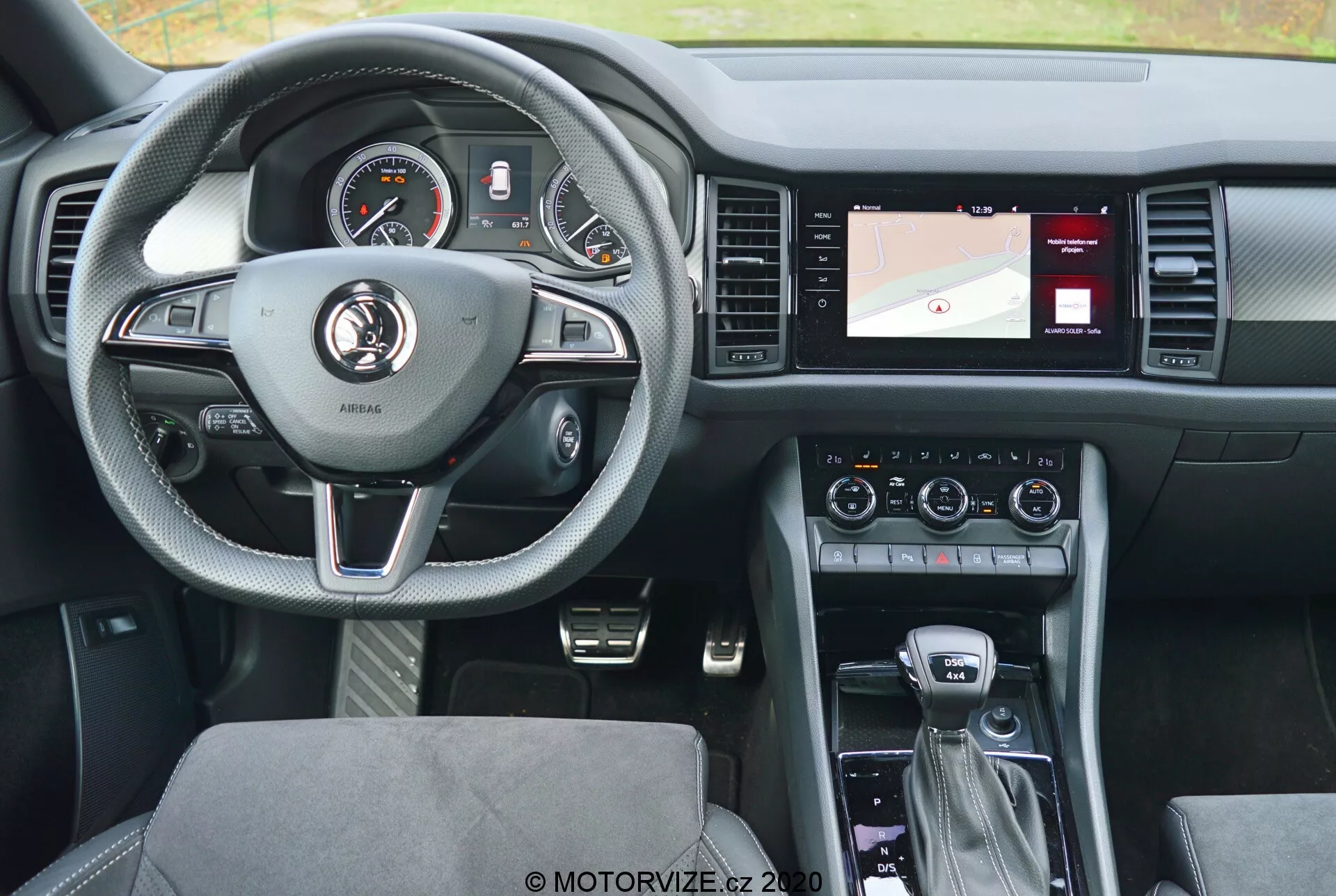 Vue intérieure d'une Skoda Kodiaq 2017 depuis la banquette arrière, mettant en avant le volant à trois branches en cuir avec accents argentés et boutons de commande pour le régulateur de vitesse, l'audio et la communication mains libres. Le tableau de bord derrière le volant comporte des cadrans analogiques et un écran numérique. La console centrale à droite comprend un écran avec une carte pour la navigation, des commandes de climatisation et des réglages audio. Le levier de vitesses suggère une transmission automatique, avec des contrôleurs supplémentaires pour le système d'infodivertissement et les modes de conduite. Le design moderne vise à équilibrer confort et technologie, avec des sièges en tissu à motifs et une disposition bien éclairée et conviviale.