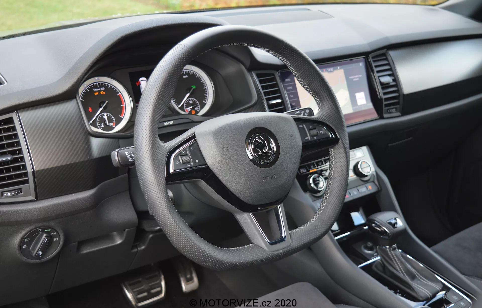 Vue intérieure d'une Skoda Kodiaq 2017 centrée sur la perspective du conducteur du tableau de bord. Le volant à trois branches, enveloppé d'un matériau semblable au cuir avec des coutures contrastantes, dispose de commandes intégrées pour l'audio, la communication mains libres et le régulateur de vitesse. Les jauges analogiques, avec le tachymètre à gauche et le compte-tours à droite, sont complétées par un écran numérique au centre. L'écran tactile d'infodivertissement, encadré par des bouches d'aération, les cadrans de contrôle du climat et le levier de vitesse sont visibles, ainsi que des matériaux texturés et des accents métalliques brossés.