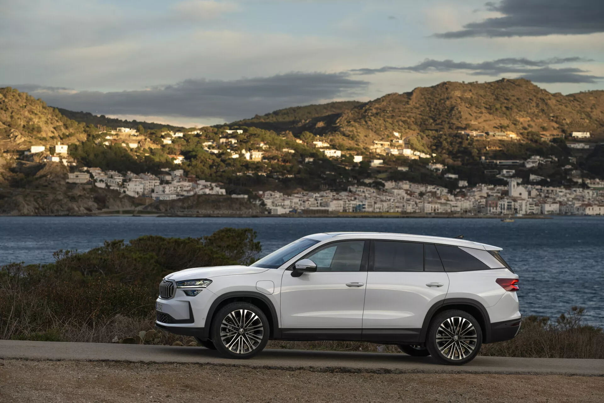 SUV moderne Skoda Kodiaq 2024 garé sur une route côtière avec un arrière-plan pittoresque de baie et de maisons sur les collines, mettant en vedette un design élégant, un empattement long, une garde au sol élevée, des lignes de carrosserie nettes, des poignées de porte affleurantes, des vitres arrière teintées, des barres de toit, de grandes roues en alliage, des phares LED, des rétroviseurs latéraux avec clignotants et un toit panoramique, le tout sur une finition extérieure blanche contrastante.