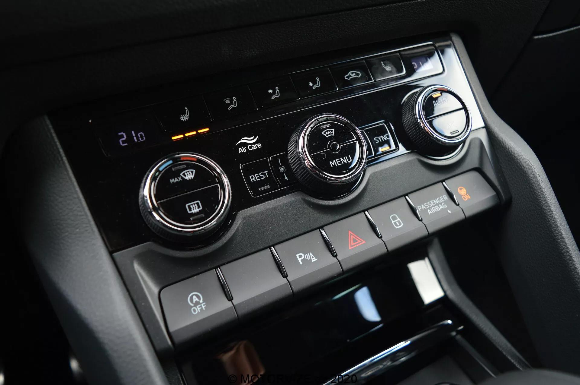 Vue rapprochée de la console centrale du Škoda Kodiaq 2017, mettant en évidence le système de contrôle climatique avec deux grands boutons de réglage de la température et un affichage numérique réglé sur 21 degrés Celsius. L'image présente également des boutons pour la climatisation, la recirculation, les dégivreurs de vitres et un bouton 'MENU'. En dessous, il y a des interrupteurs à bascule pour les capteurs de stationnement, la désactivation de l'auto start/stop et les feux de détresse.