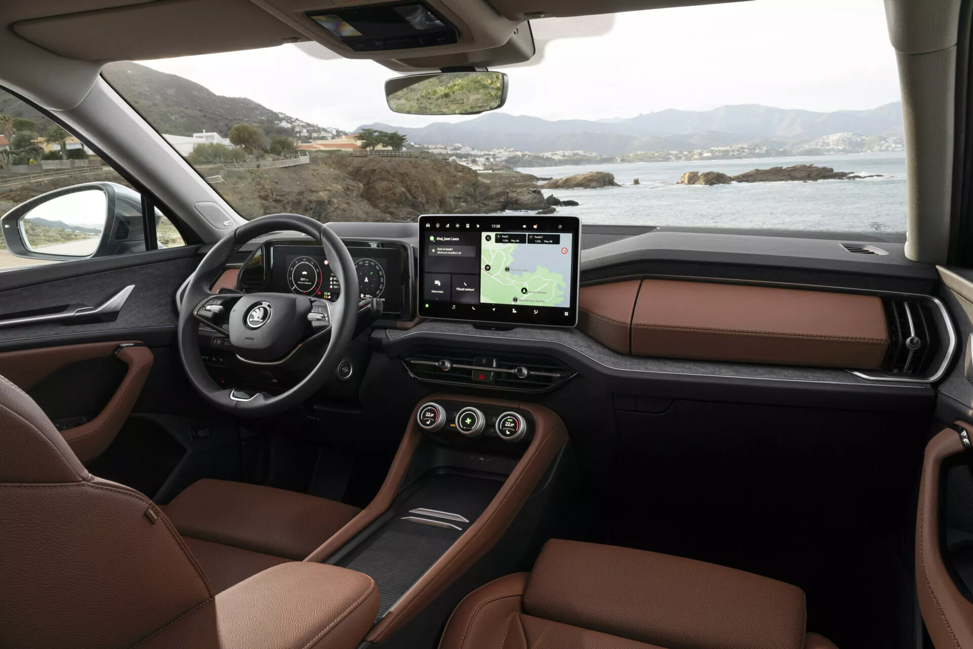 Vue intérieure du Skoda Kodiaq 2024 du point de vue du passager montrant le tableau de bord, le volant multifonction avec logo, le cluster d'instrumentation numérique, le grand système d'infodivertissement tactile avec GPS, les sièges en cuir marron luxueux et la conception élégante de la console centrale, situé contre un paysage côtier serein à l'extérieur des fenêtres