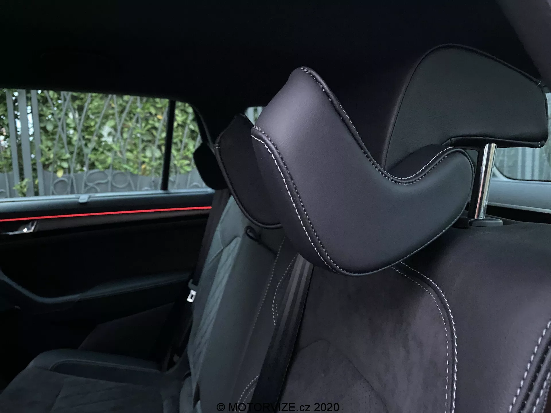 Vue intérieure d'une Skoda Kodiaq 2017 se concentrant sur un siège de voiture. La prise de vue en angle met en lumière la partie appuie-tête, rembourrée de matériau noir, probablement en cuir ou en synthétique de haute qualité, avec un contour distinct pour le confort. Les coutures précises avec un fil contrastant ajoutent des accents luxueux. À l'arrière-plan, un éclairage d'ambiance rouge horizontal souligne les éléments de design automobile moderne. Les commodités visibles pour les passagers arrière comprennent des ceintures de sécurité et peut-être un accoudoir central, indiquant la deuxième rangée de sièges.