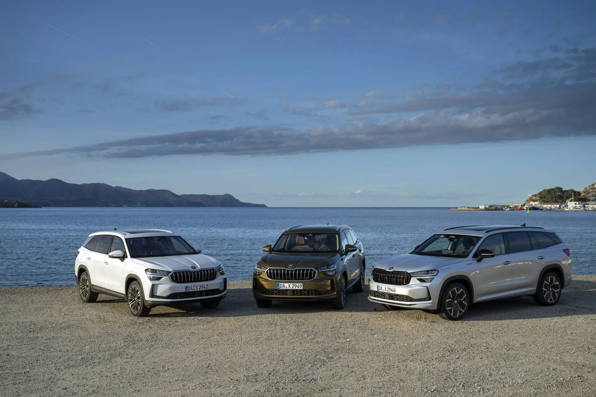 Gamme de SUV Skoda Kodiaq 2024 en blanc et en couleurs foncées, vue de côté avant, garés sur un front de mer avec des montagnes et un ciel partiellement nuageux en arrière-plan