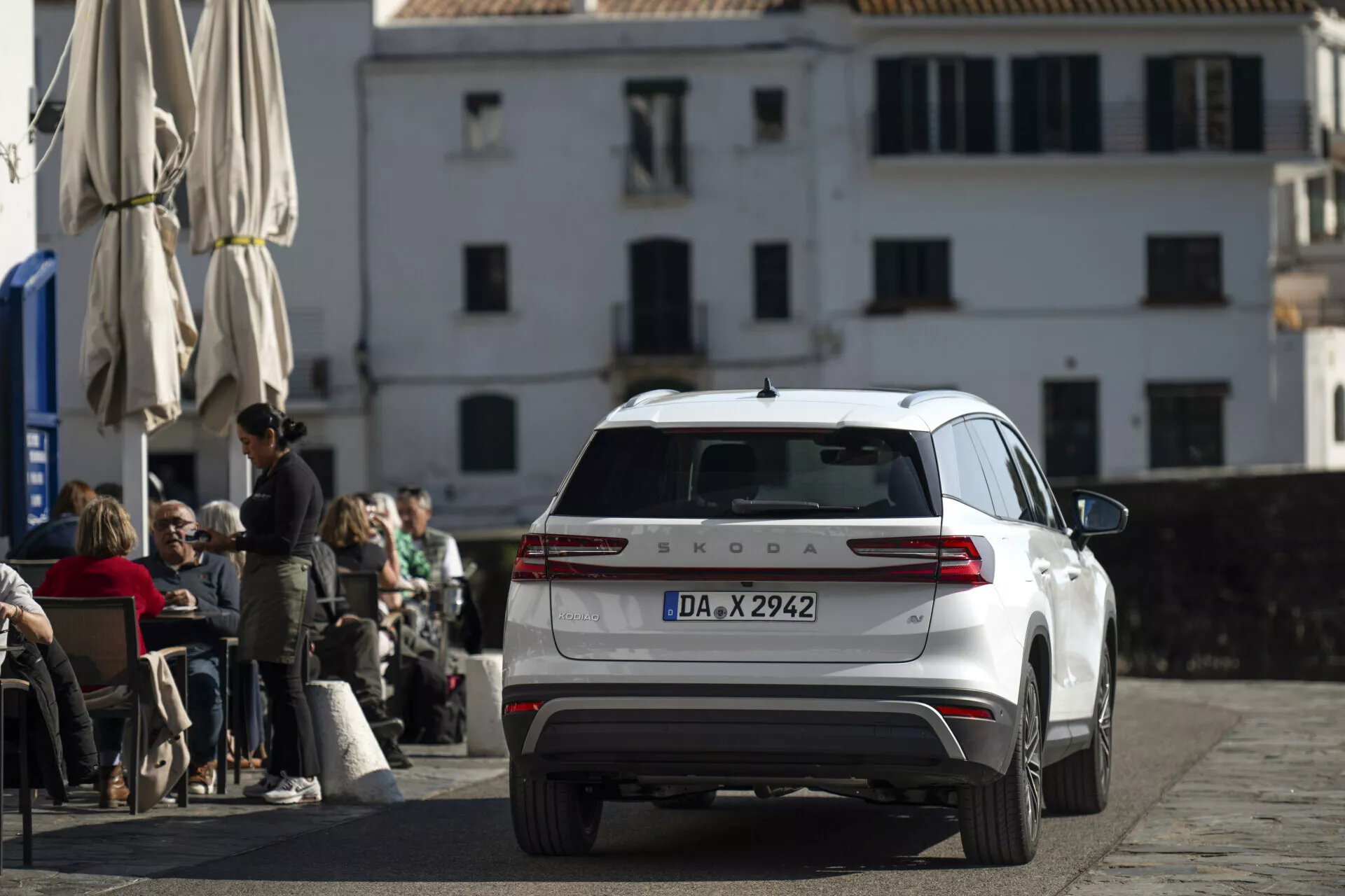 Škoda Kodiaq blanche 2024 photographiée depuis un angle arrière et légèrement surélevé, montrant l'arrière complet, les feux arrière, le couvercle du coffre, la fenêtre arrière avec essuie-glace et le toit avec antenne et barres, stationnée sur une rue pavée devant un café avec une architecture européenne classique en arrière-plan.