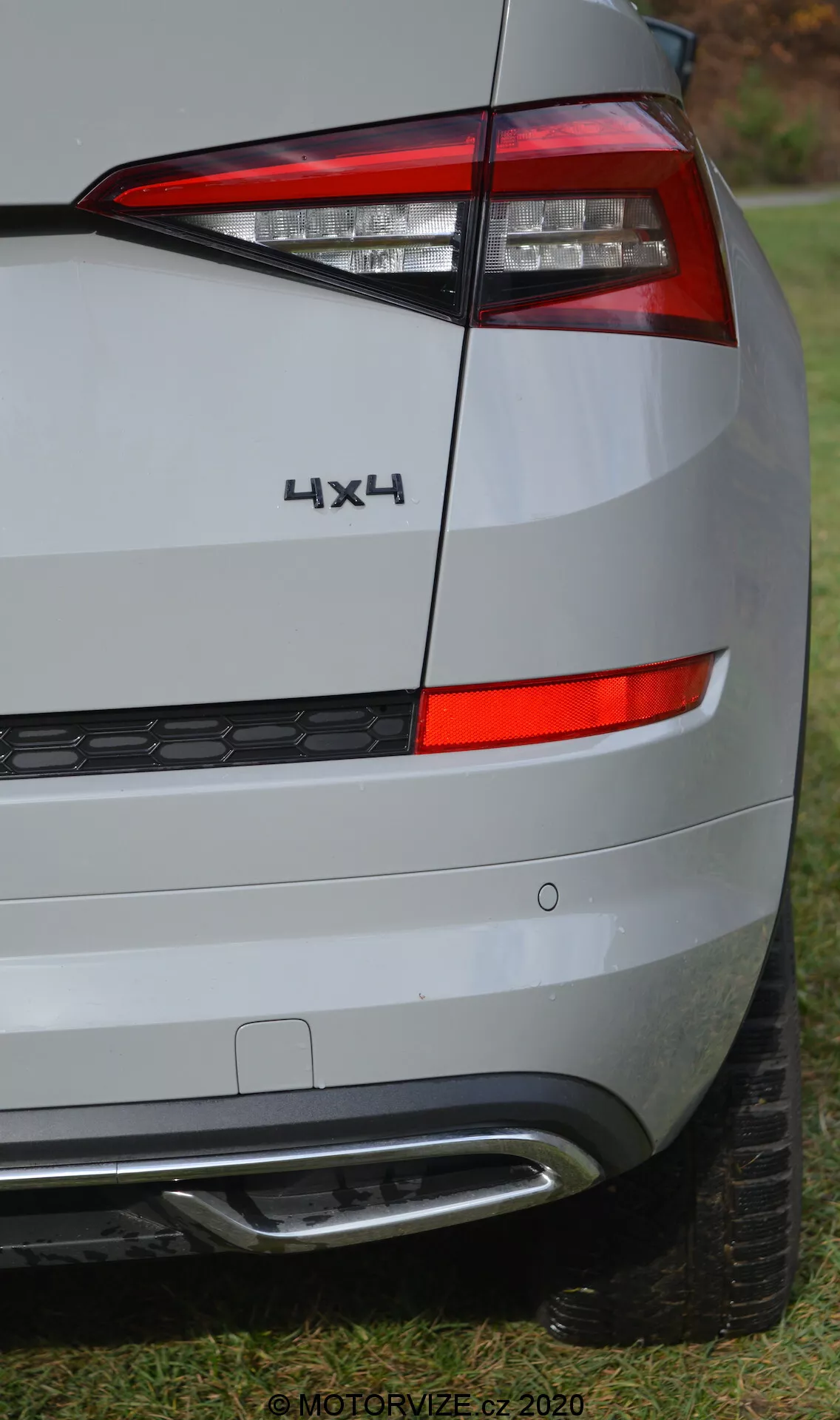 Vue partielle rapprochée de l'angle arrière d'une Skoda Kodiaq 2017, se concentrant sur le design de l'éclairage arrière, les badges et le pare-chocs. L'angle de la prise de vue met en évidence le côté droit de l'arrière du véhicule, montrant un ensemble de feux arrière horizontalement agencés avec un design angulaire net qui enveloppe le côté. Sous le feu arrière, on peut voir le panneau arrière et le pare-chocs, de couleur argentée ou grise claire, reflétant un environnement atténué. Le pare-chocs comprend une section inférieure en plastique noir avec un motif en nid d'abeille et un élément en argent ou chromé, probablement lié à l'assistance au stationnement ou au design. Un badge '4X4' au-dessus du feu arrière indique une transmission intégrale. L'image haute résolution révèle les textures de surface détaillées, une partie de la bande de roulement étant visible en bas, positionnée sur de l'herbe. Le filigrane '© MOTORVIZE.cz 2020' indique que l'image est destinée à une publication automobile tchèque.