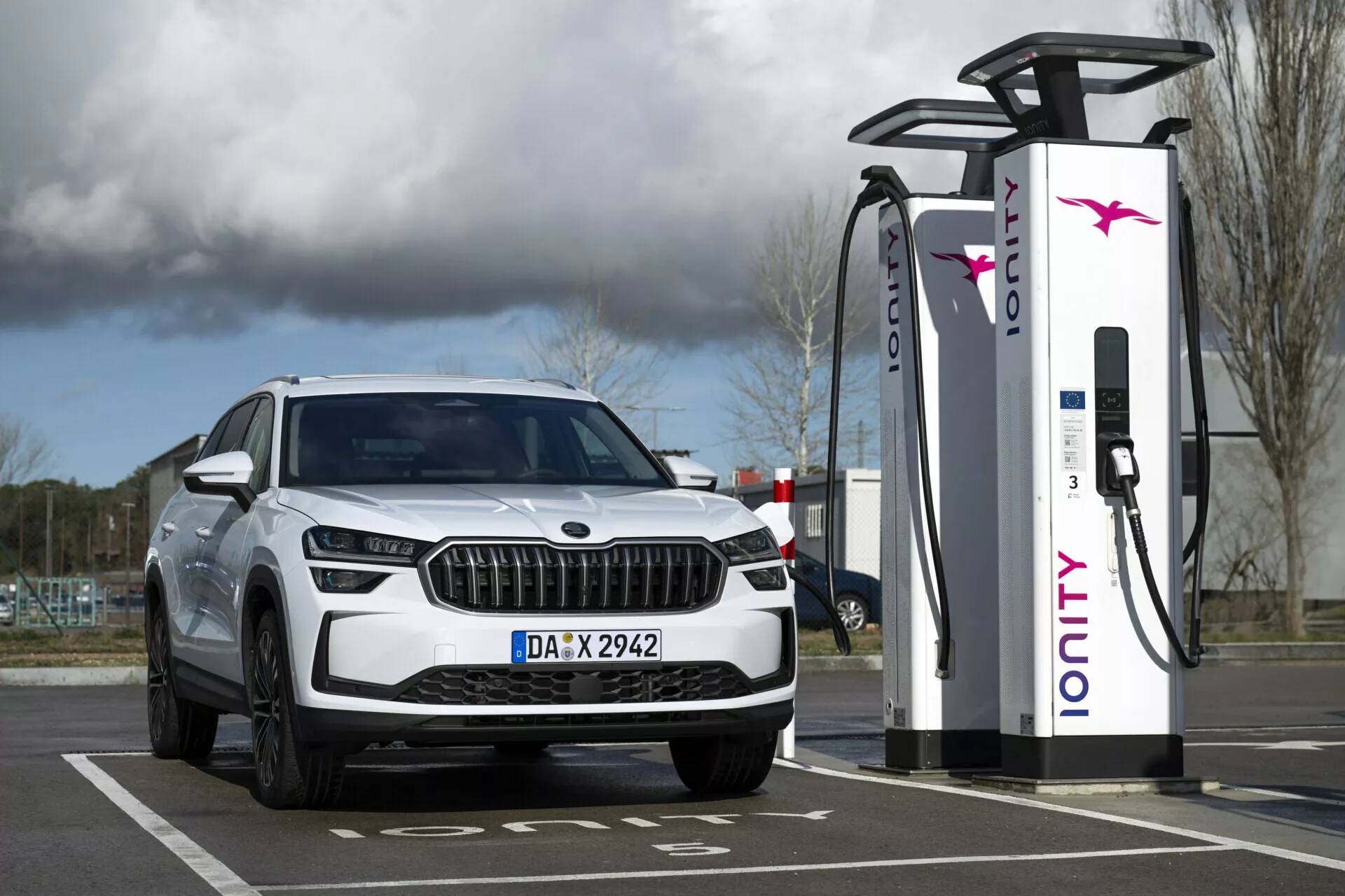 SUV blanc Skoda Kodiaq 2024 garé à une station de recharge électrique, vue de l'avant avec phares LED et feux de jour, soulignant ses capacités électriques ou hybrides et son design de grille luxueux.