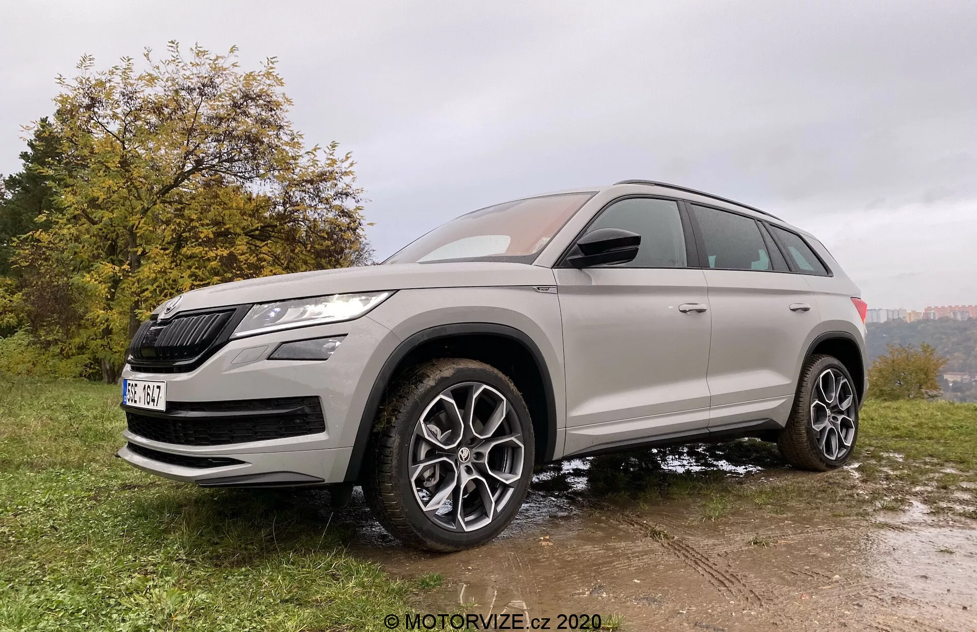 Vue trois-quarts avant d'un SUV Škoda Kodiaq 2017 prise d'un angle légèrement bas, soulignant sa hauteur et sa posture. Le véhicule est garé sur une surface de gazon et de boue irrégulière, mettant en valeur ses capacités tout-terrain. Le Škoda Kodiaq présente une calandre proéminente et des phares LED affilés. Le design minutieux comprend de grandes roues en alliage à plusieurs rayons, une grande garde au sol et des pneus toutes saisons. La carrosserie est peinte dans une couleur métallique neutre avec des vitres teintées, et des accents chromés rehaussent son attrait premium. Les fonctionnalités modernes se remarquent dans les rétroviseurs latéraux de la couleur de la carrosserie avec des clignotants intégrés et peut-être des systèmes d'avertissement d'angle mort.