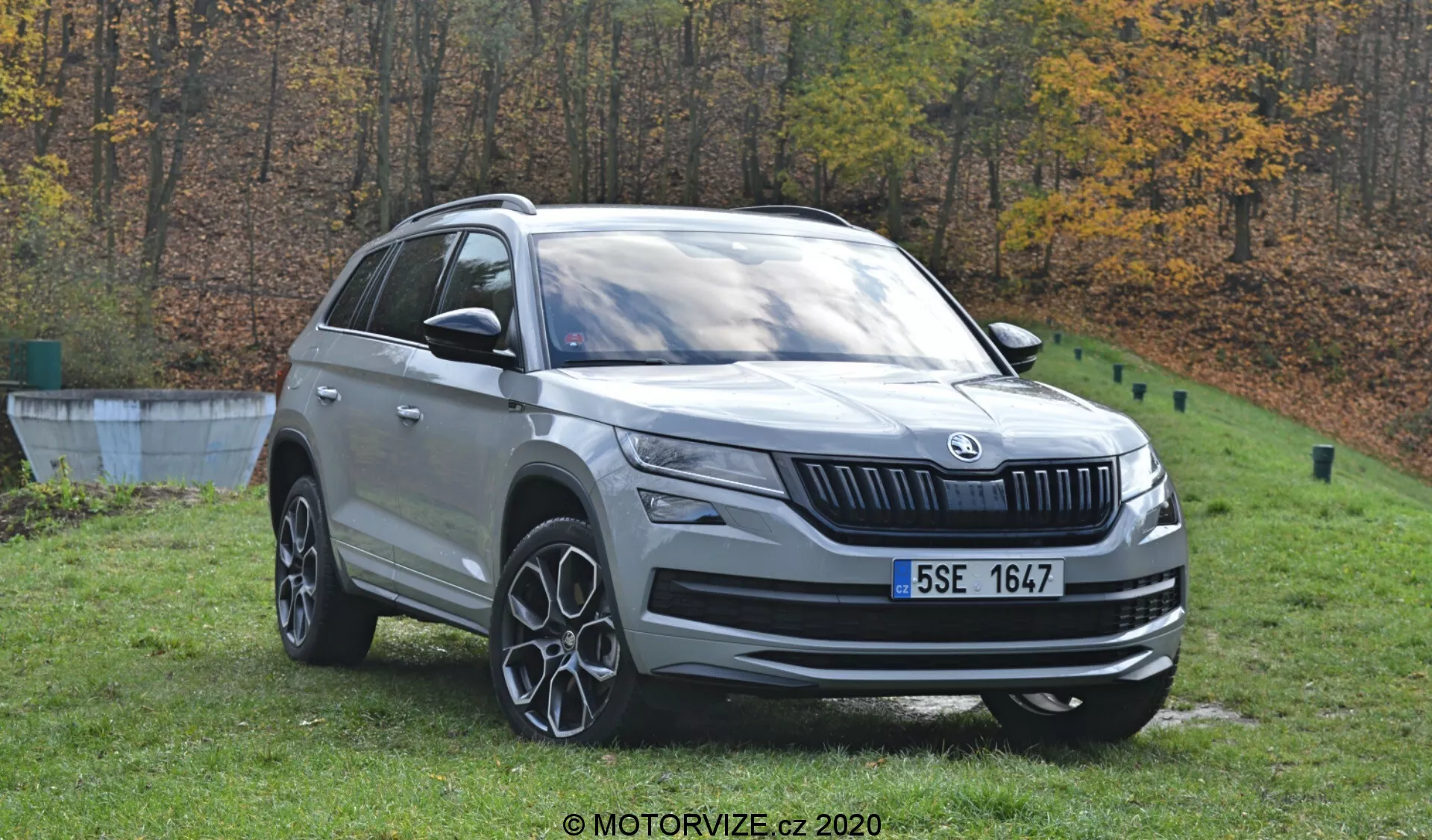 Une vue détaillée de la Škoda Kodiaq 2017, un SUV de couleur argent, placé en extérieur avec en arrière-plan des arbres en automne. L'image prise en plein jour capture le véhicule sous un angle trois-quarts avant, montrant son profil avant et latéral. Des éléments de design distinctifs sont visibles, notamment l'emblème de la marque sur la calandre avant, les phares à LED, les phares antibrouillard, les roues en alliage multi-rayons, les rails de toit et les rétroviseurs latéraux avec clignotants intégrés. La voiture est positionnée sur l'herbe près d'un bord en béton, ce qui suggère un environnement de type parc.